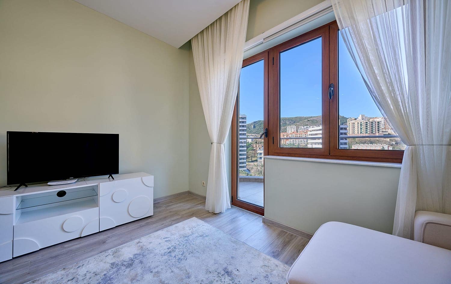 Living room in an apartment in Becici, Montenegro: bright interior, TV area, panoramic window with a view of the mountains and the city.