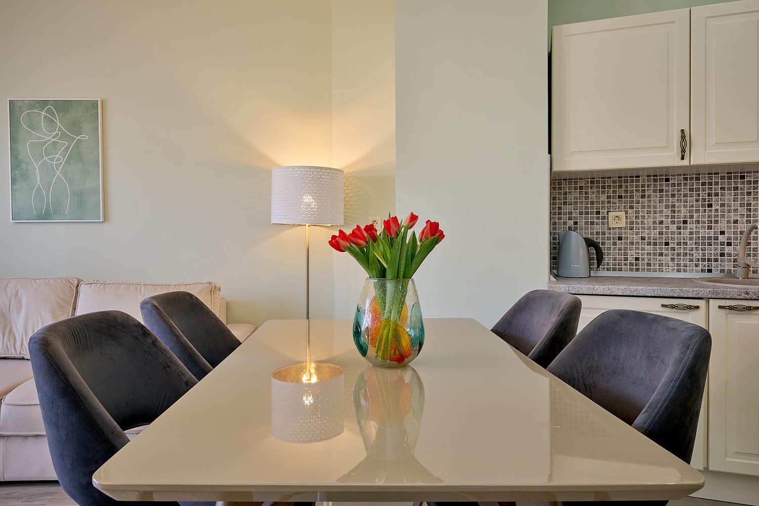 Dining area in a Bečići apartment, Montenegro: light-colored table, grey chairs, kitchen cabinetry, and tulip decor.