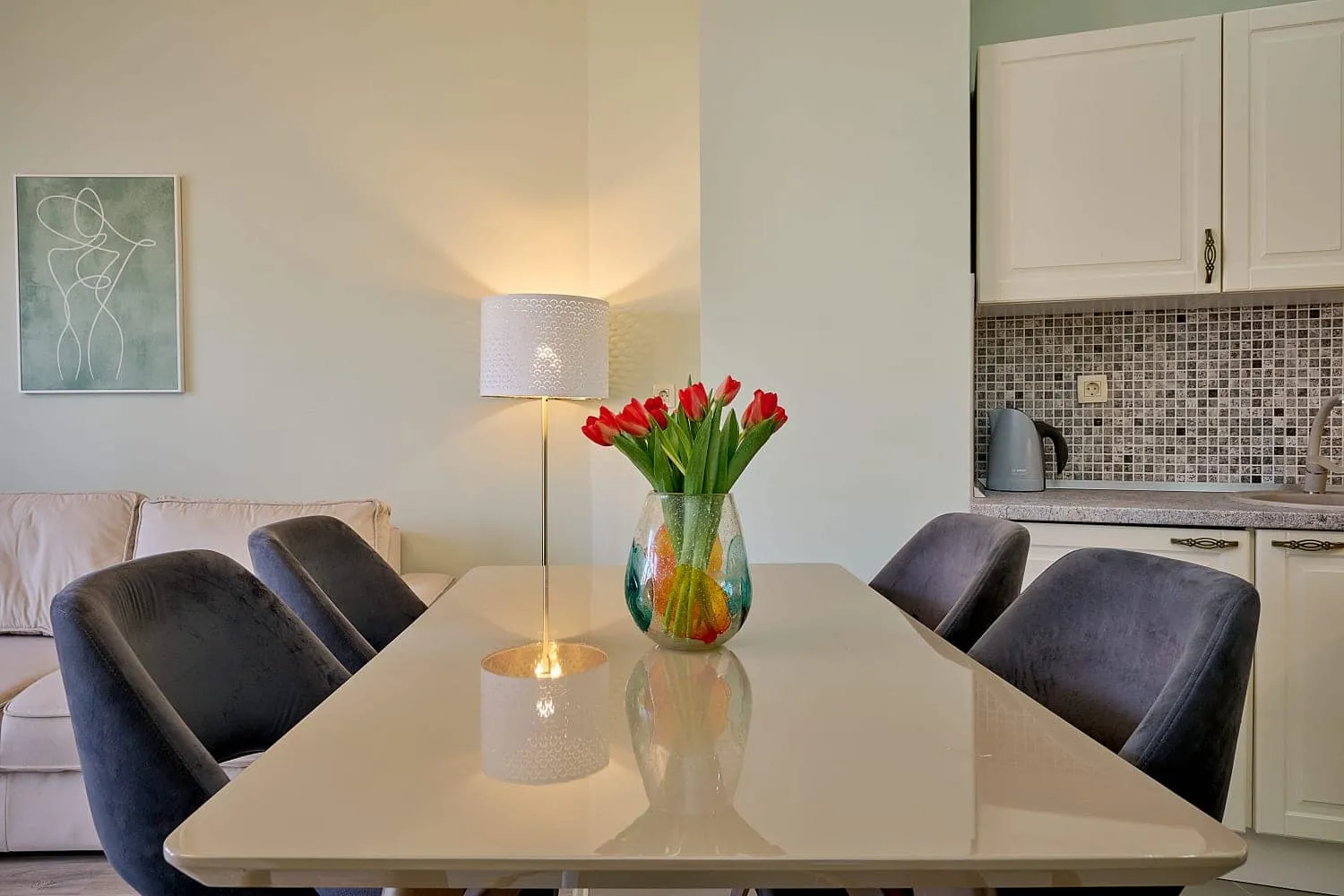 Dining table with grey chairs, sofa, and kitchen area with white cabinets visible in the background.
