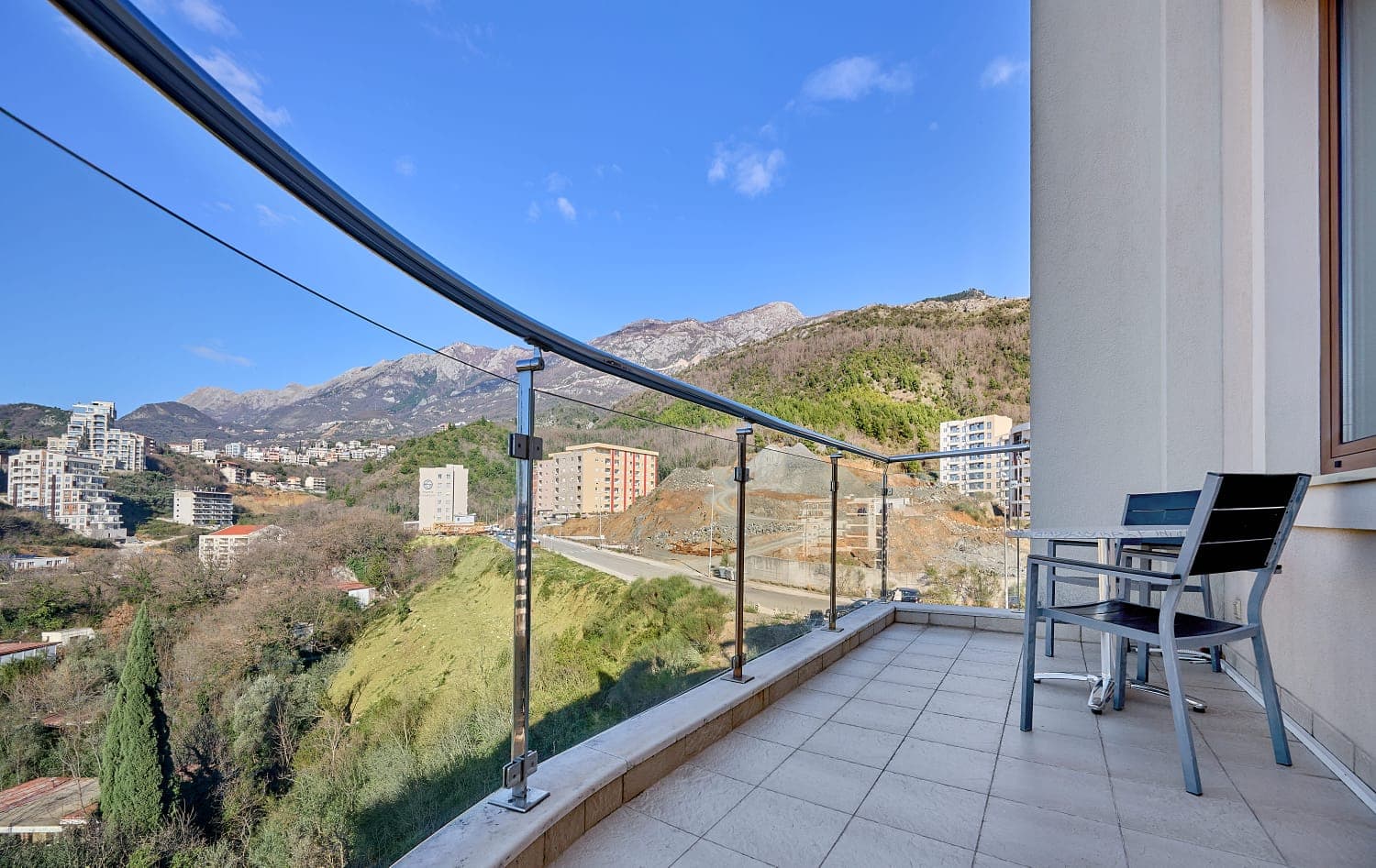 Balcony with furniture and a panoramic view of the mountains and the town of Bečići, Montenegro. Modern glass and metal railing.