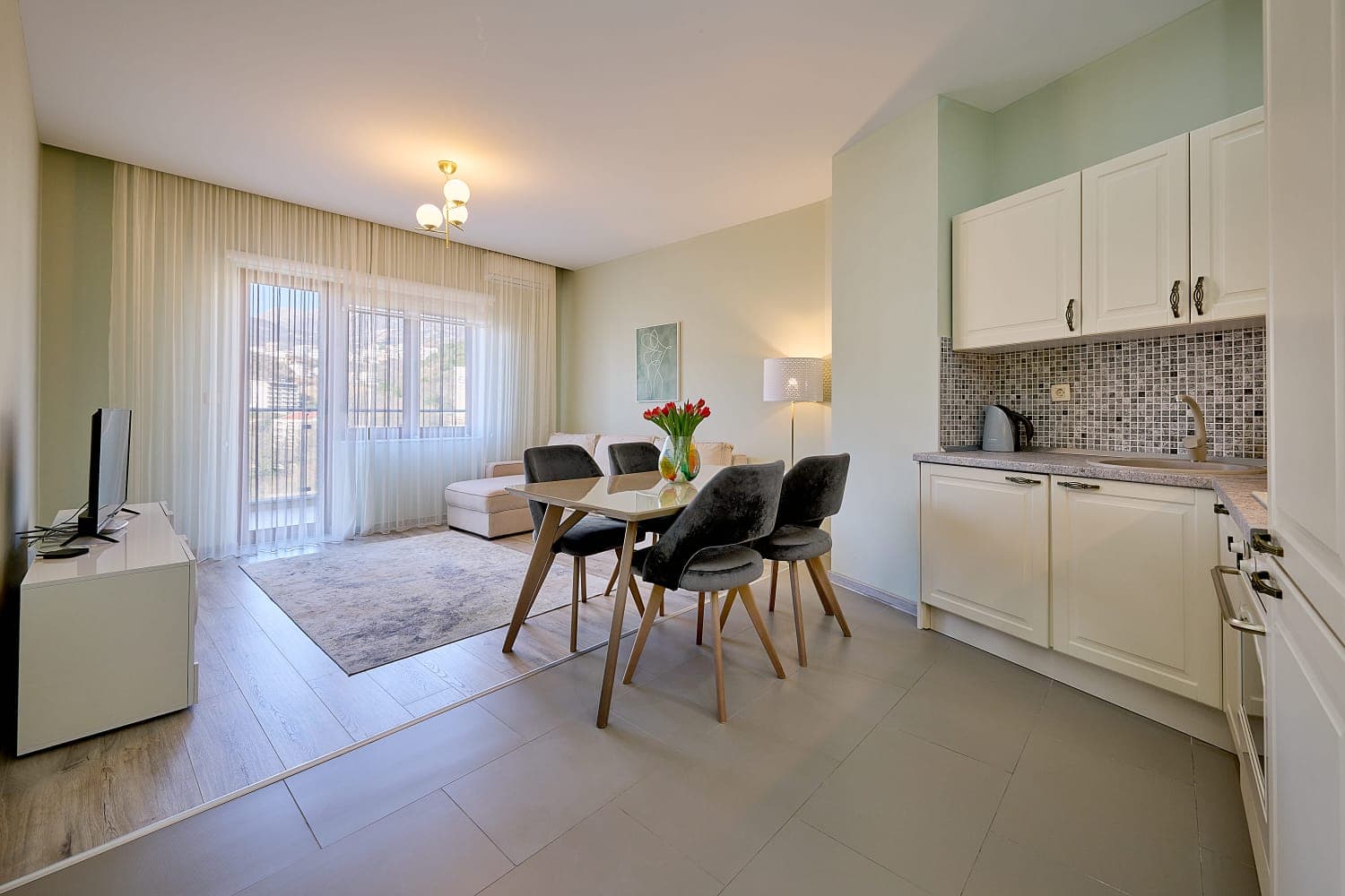 Living room with kitchen in an apartment in Becici, Montenegro: light-colored furniture, dining area, balcony access, and panoramic windows.