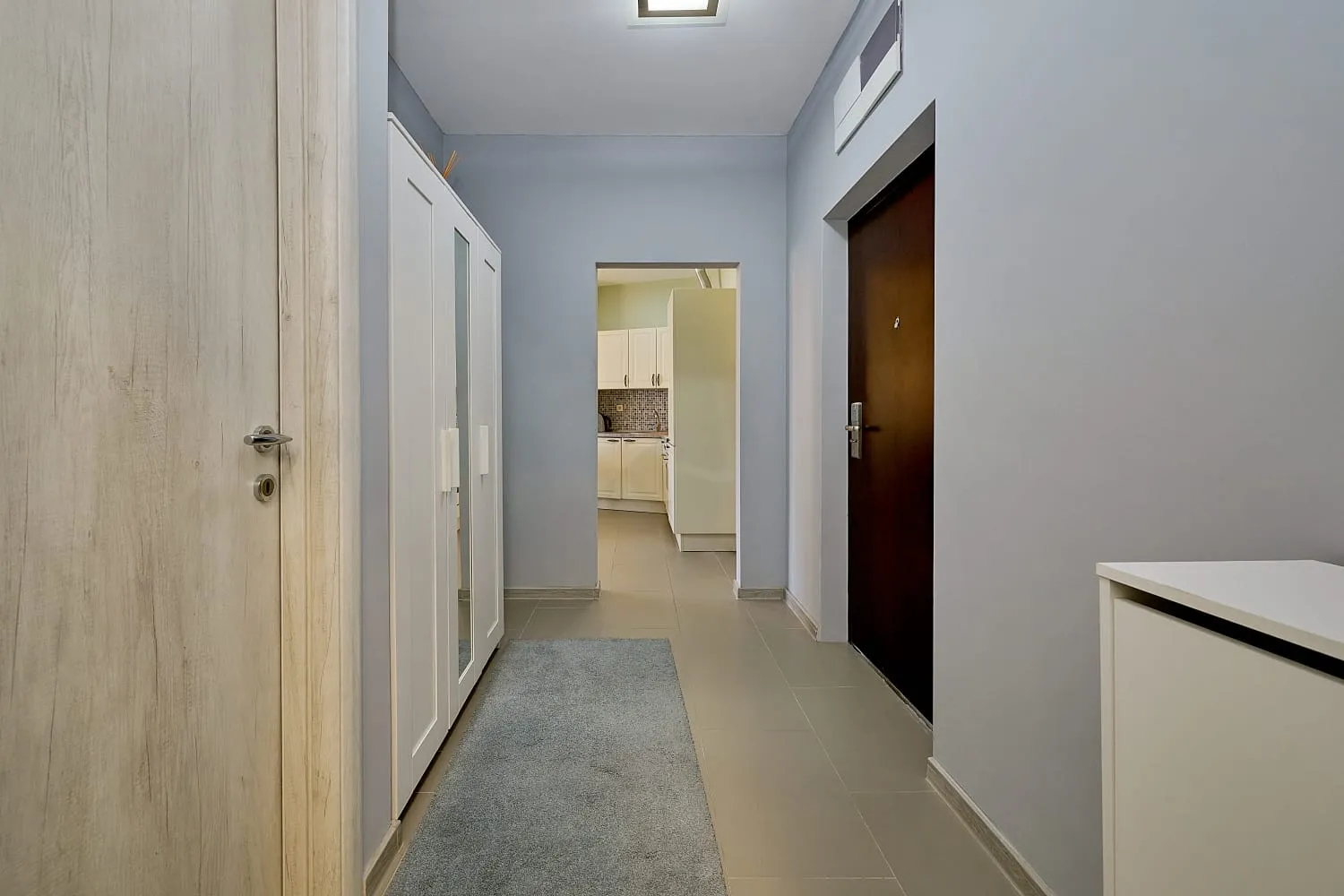 Hallway with white wardrobe, grey rug, and dark entrance door; kitchen visible through doorway.