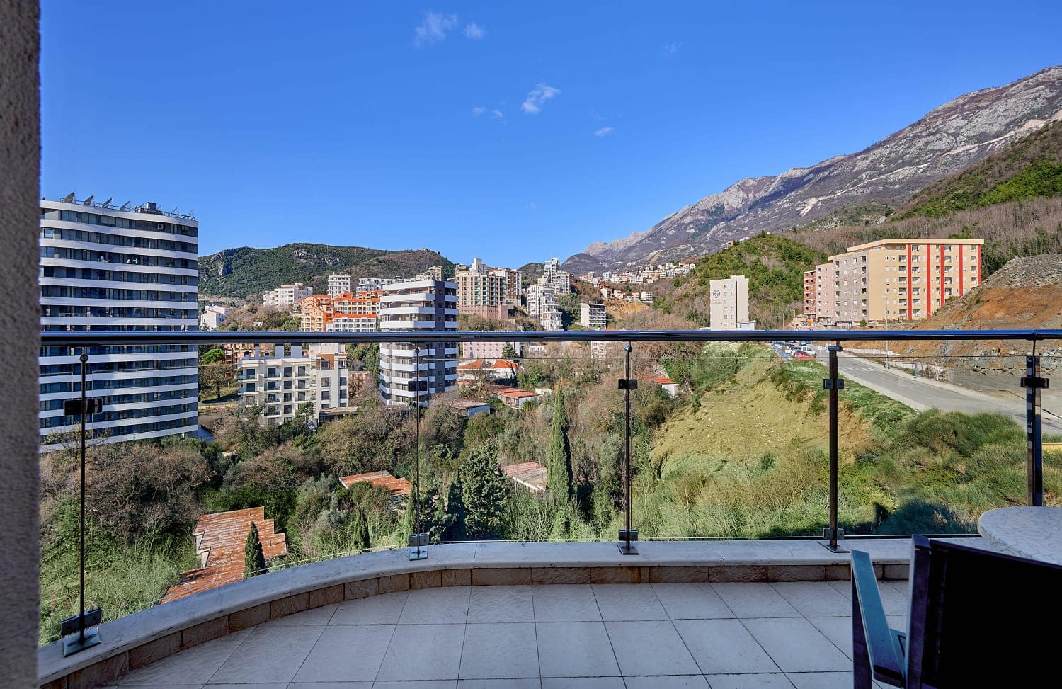 View from the apartment balcony in Bečići, Montenegro: a panorama of mountains, modern architecture, and green hills on a sunny day.