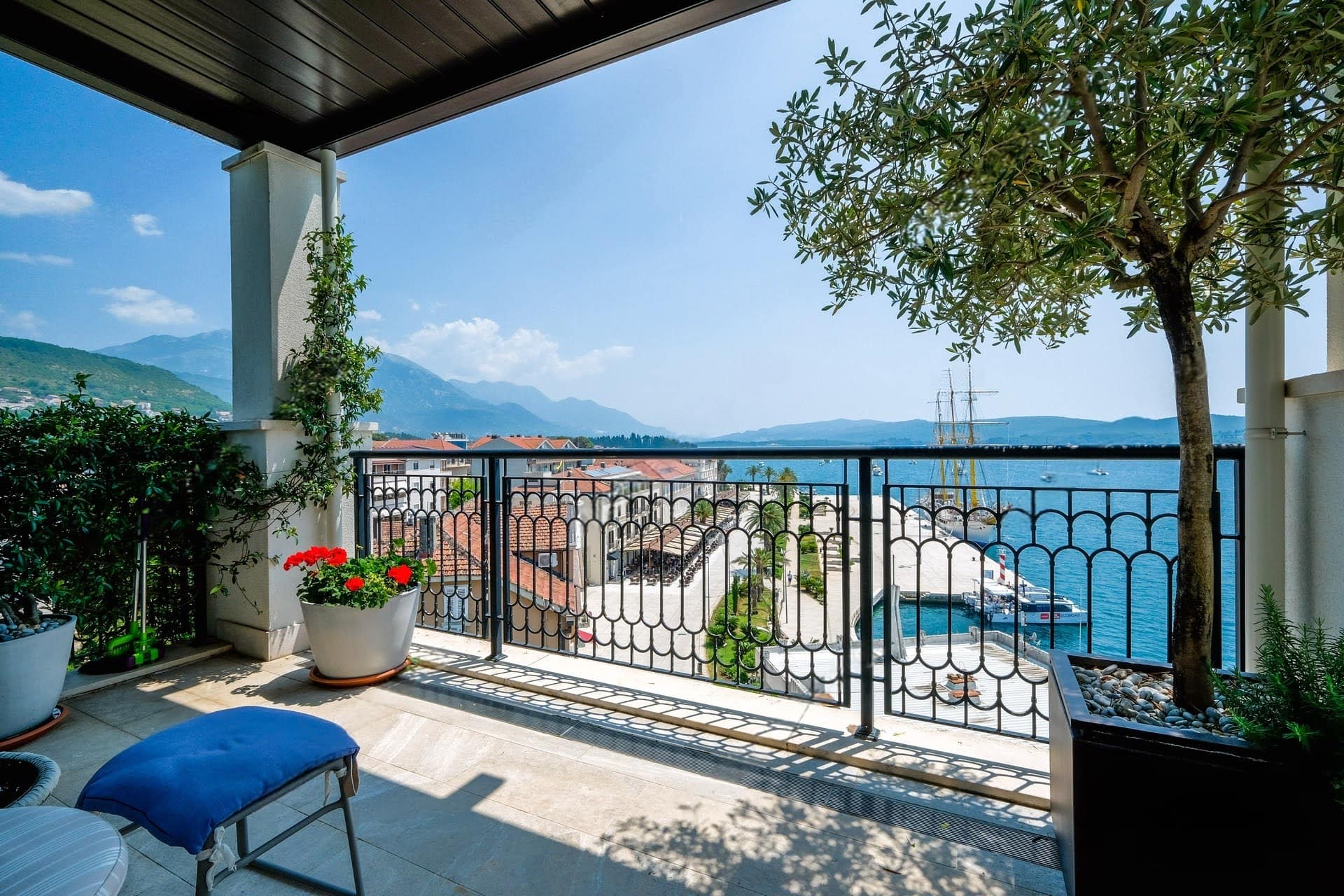 Apartment terrace in Tivat, Montenegro: sea, promenade, and mountain views, wrought-iron railing, olive tree in a planter.