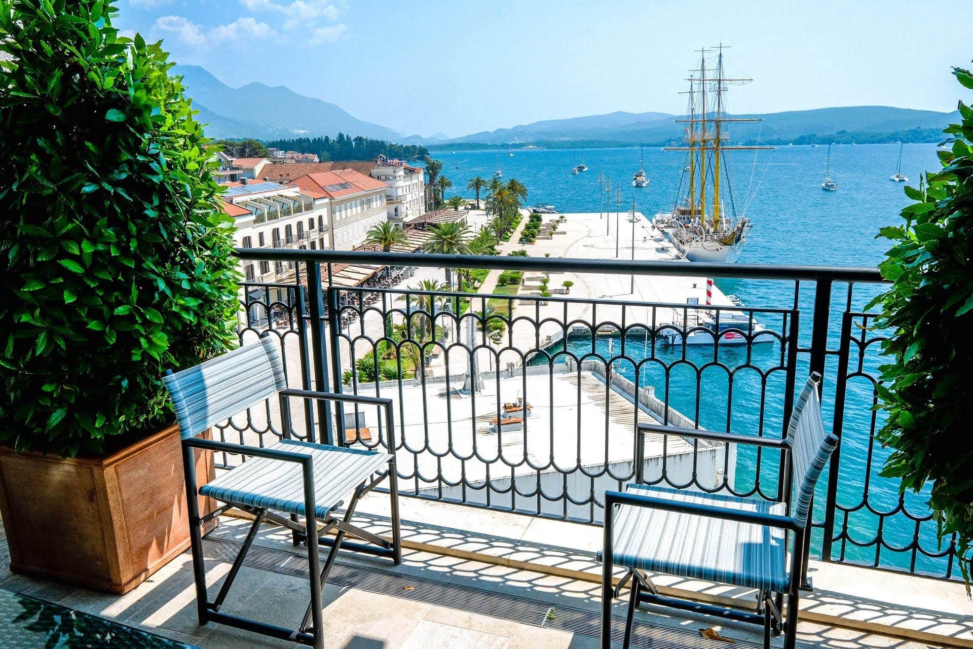 View from the apartment balcony in Tivat, Montenegro: outdoor seating, the Porto Montenegro promenade, and a sailboat at sea.