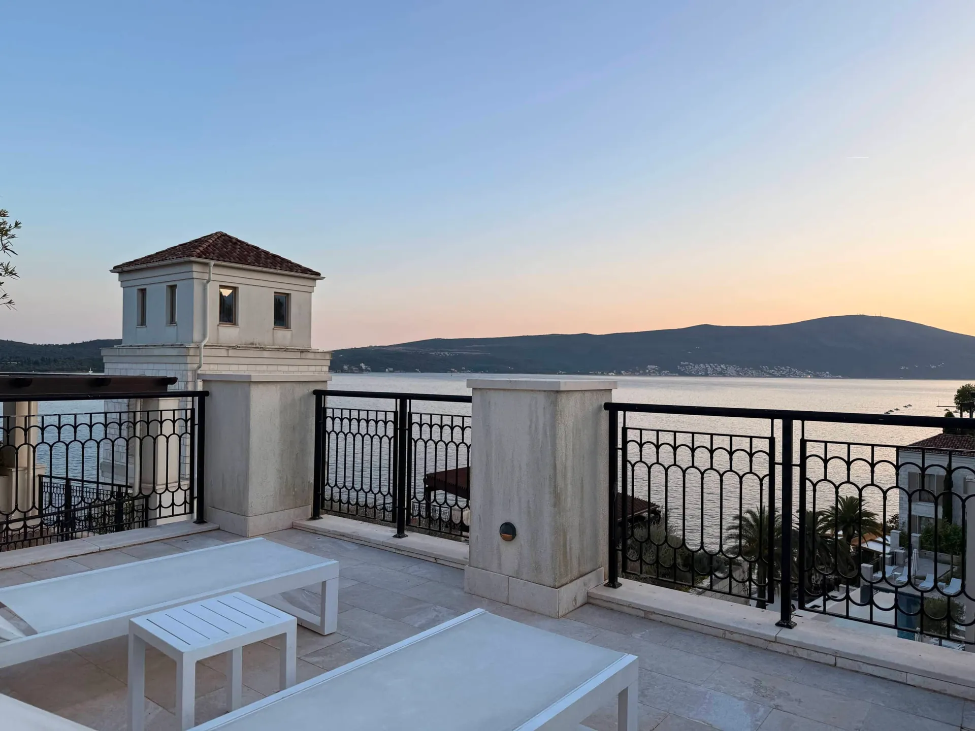 Terrace with white benches and black iron railing overlooking the sea and mountains at sunset.