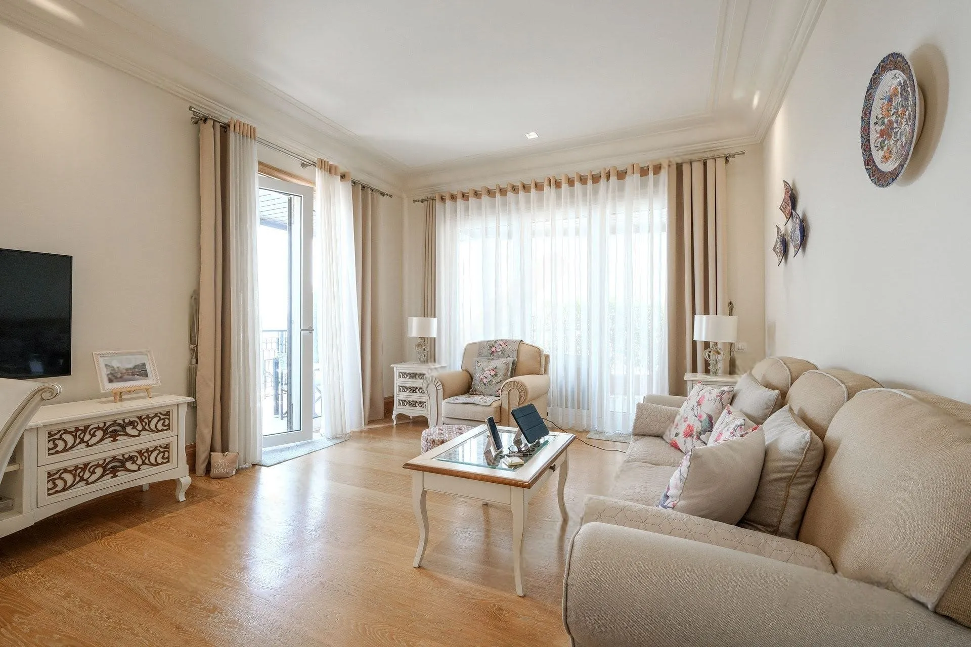 Bright room with beige sofa, coffee table, TV stand, and balcony visible behind sheer curtains.