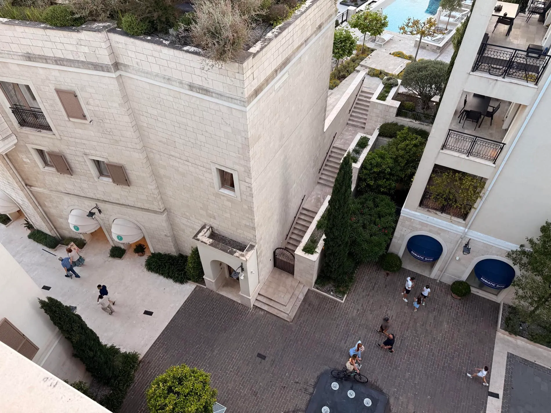 High-angle view of a stone building, brick courtyard with people, stairs and greenery.