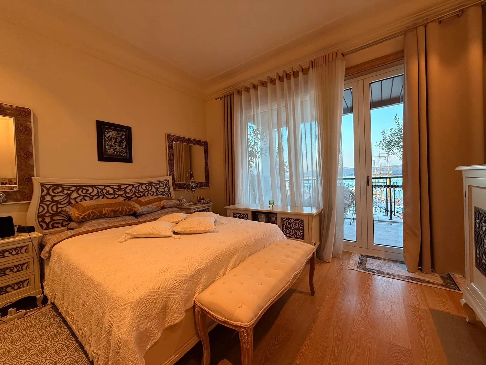 Bedroom featuring a large bed with carved headboard, upholstered bench, and glass doors leading to a balcony.