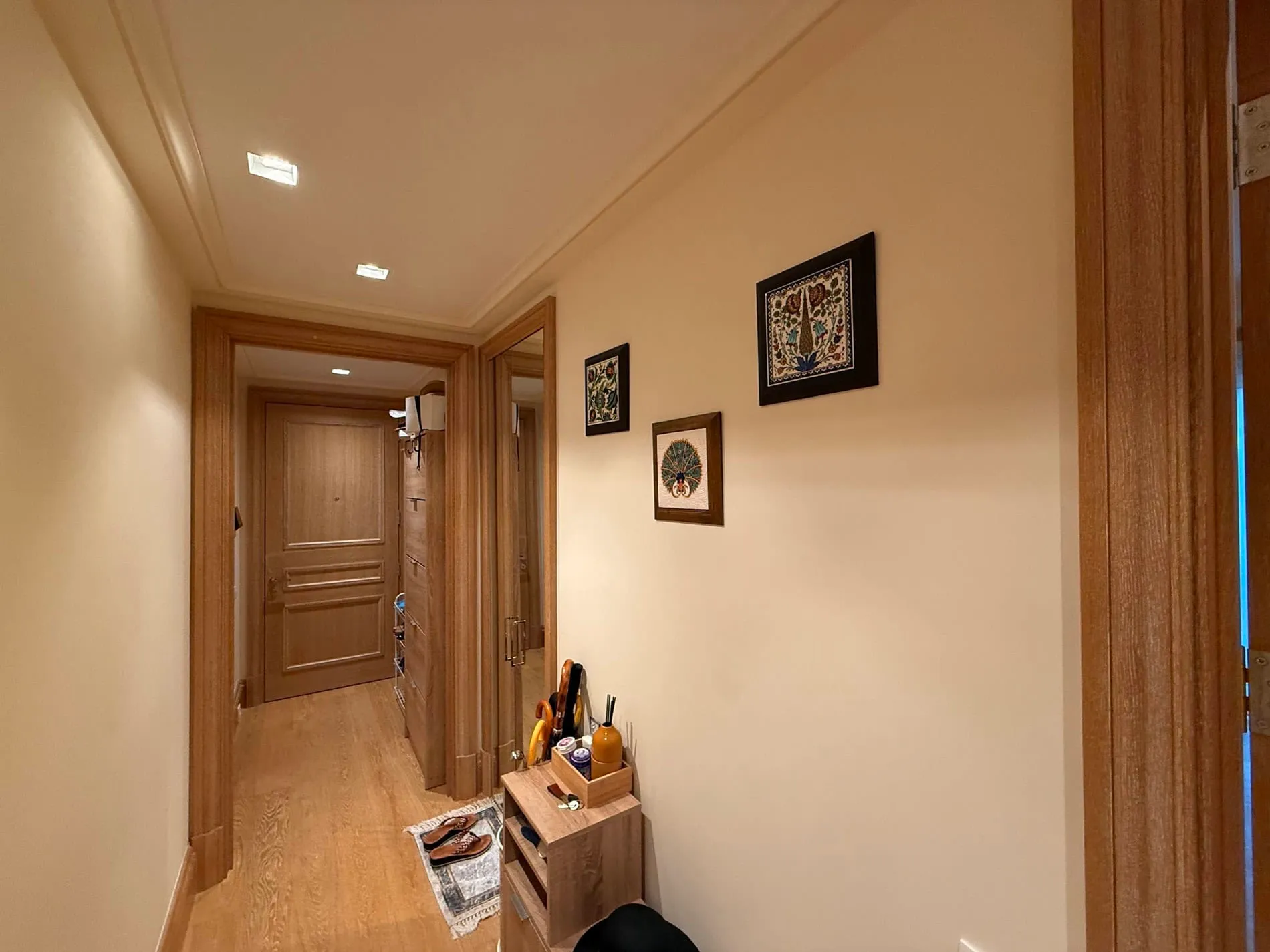 Interior hallway with light wood flooring, matching door frames, three framed artworks, and a small console holding shoes and umbrellas.