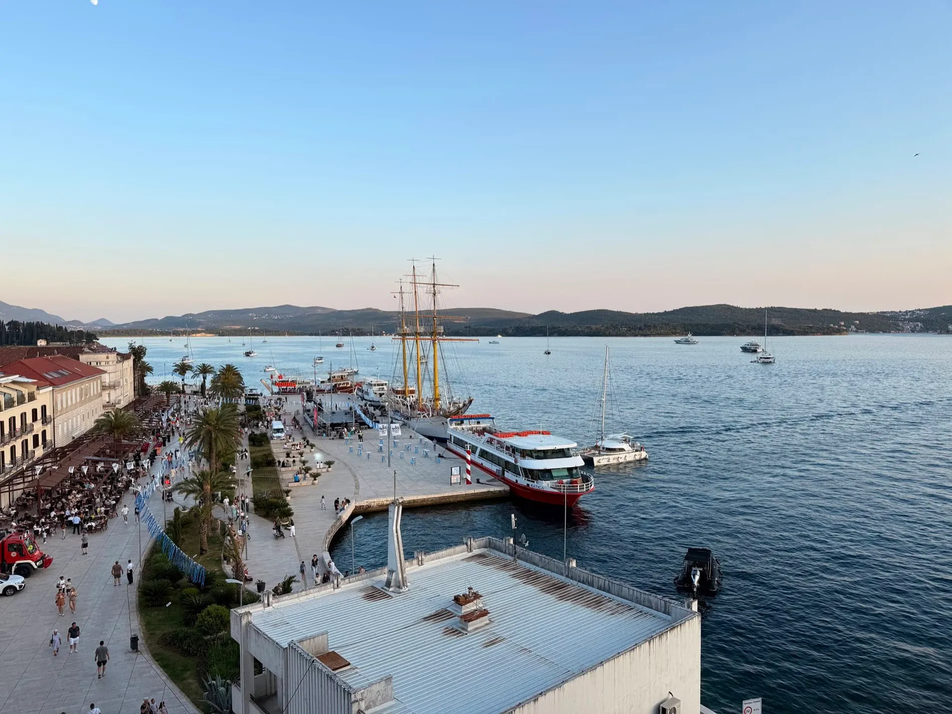 Panoramic view of a seaside port, crowded promenade, docked ships including a tall ship, and distant mountains.