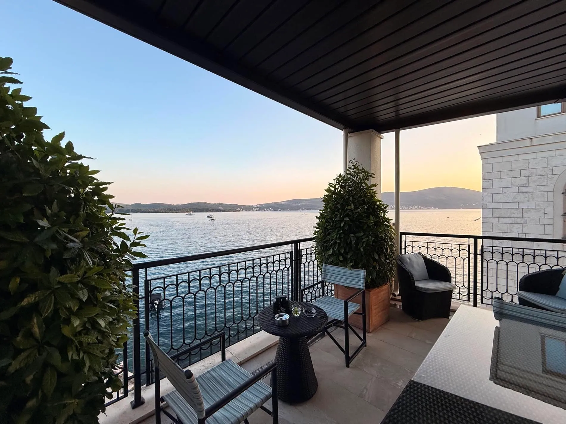 Balcony with black metal railing, outdoor furniture, and a scenic view of the sea and mountains at sunset.
