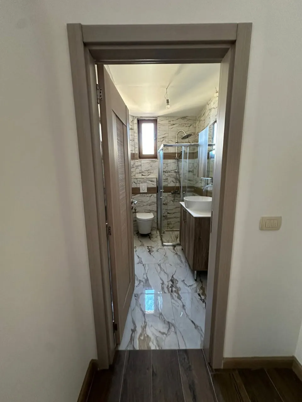 View through a doorway into a bathroom featuring marble tiles, a glass shower enclosure, toilet, and vessel sink on a wooden vanity.