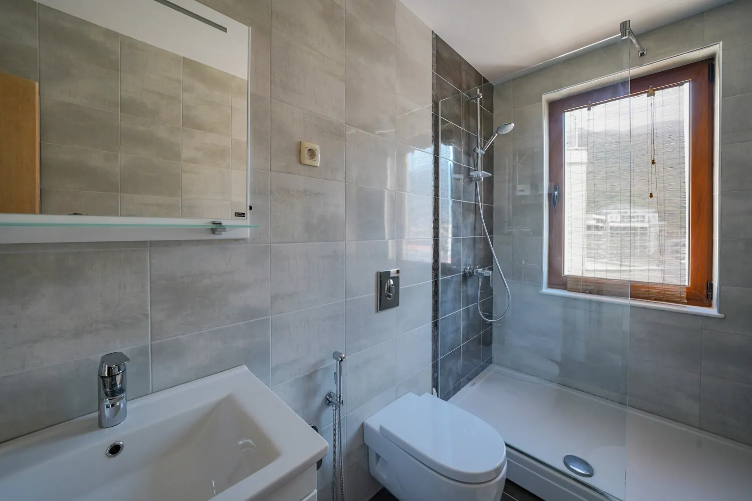 Modern bathroom with grey tiles, white sink, wall-mounted toilet, and a shower area enclosed by a glass partition near a window.