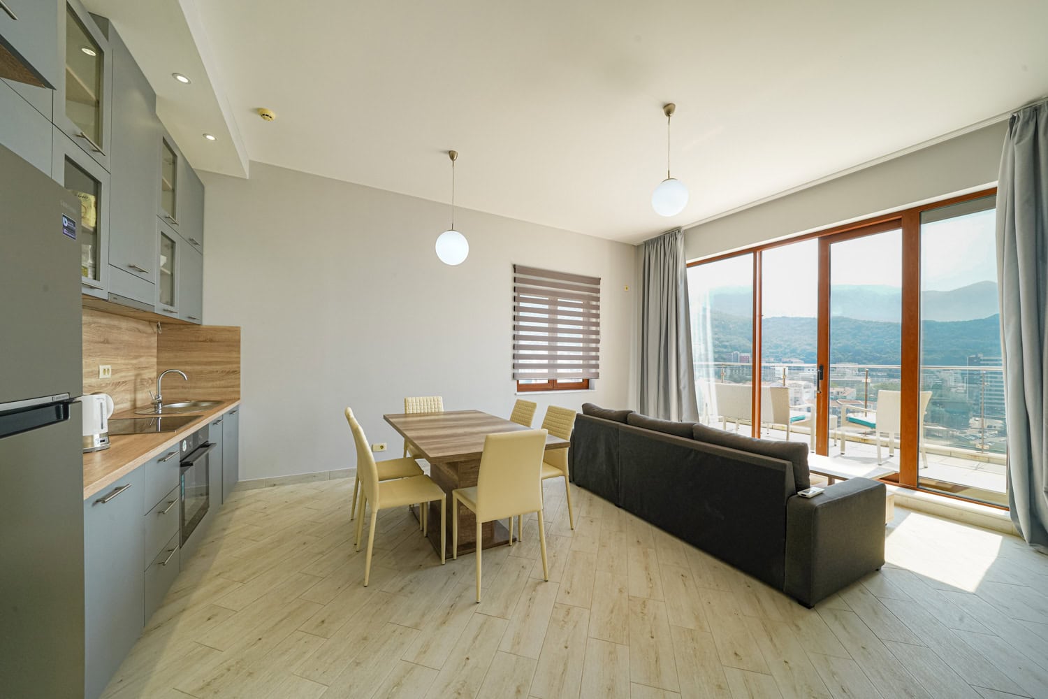 Modern living room with kitchen in an apartment in Bečići, Montenegro: grey cabinetry, dining area, and mountain views from the terrace.