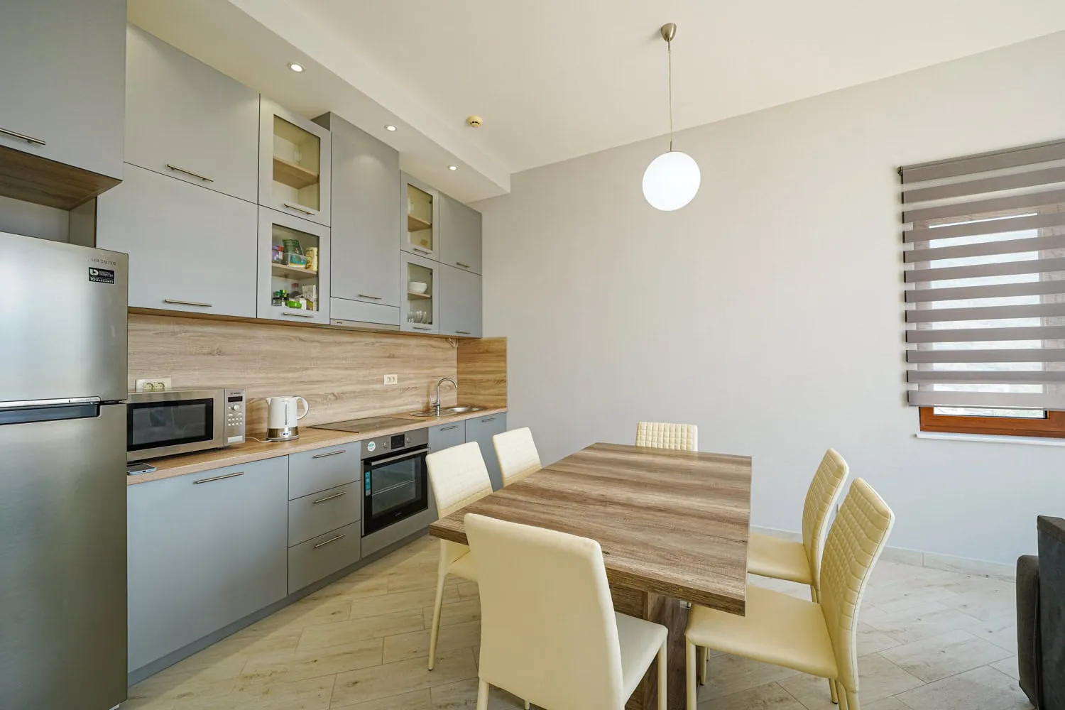 Modern kitchen with grey cabinets, wooden dining table, light chairs, and appliances like fridge and oven.