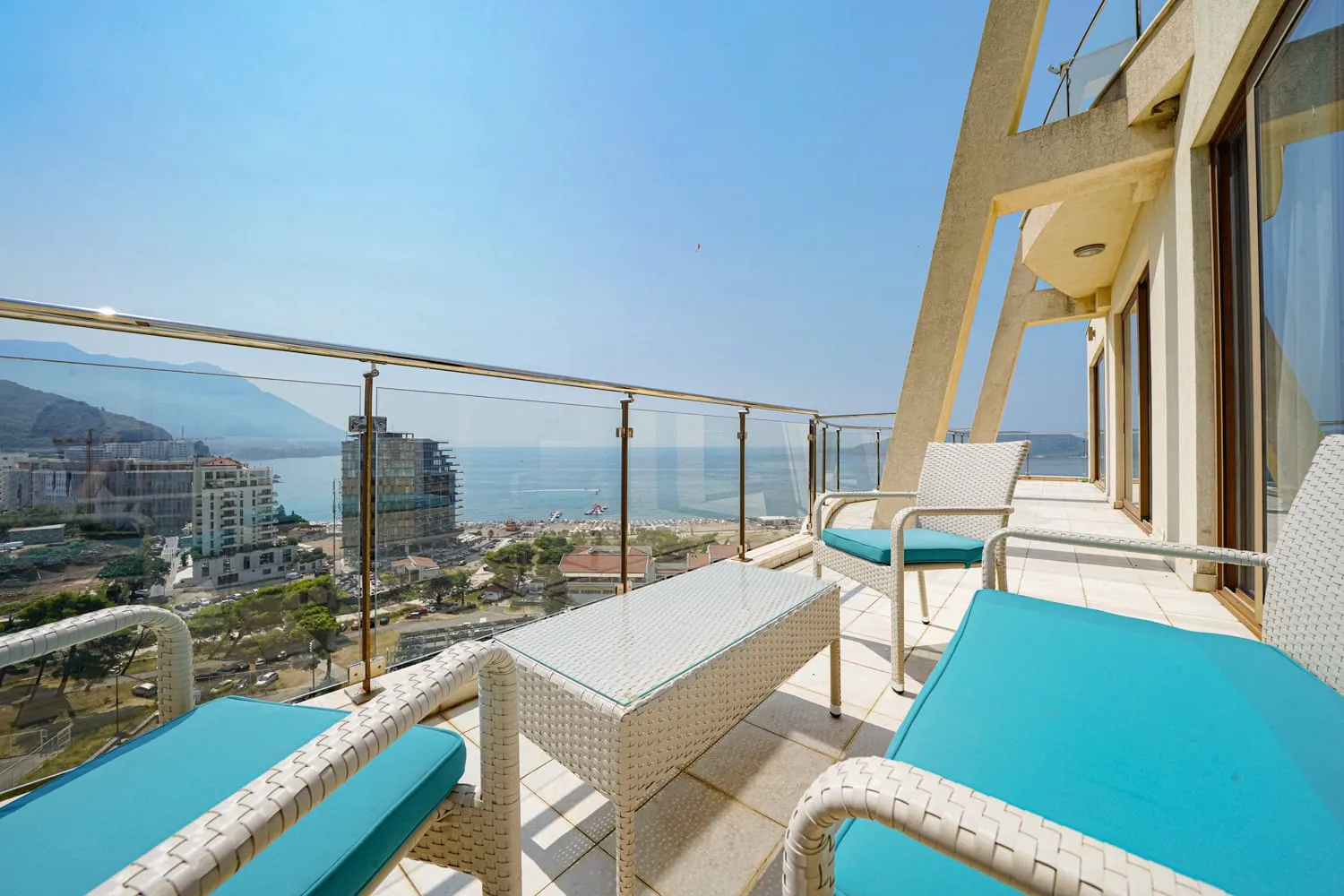 Balcony with white wicker furniture and turquoise cushions overlooking the sea and city behind a glass railing.