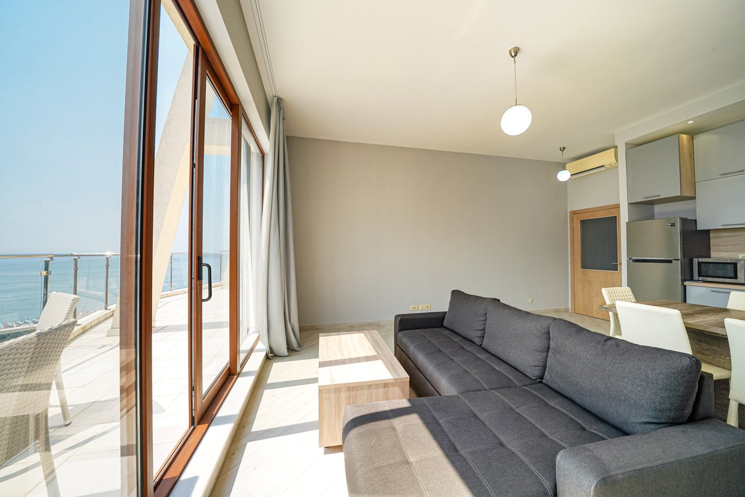 Living room with a panoramic sea view in an apartment in Bečići, Montenegro: gray sofa, kitchen, and access to the terrace.