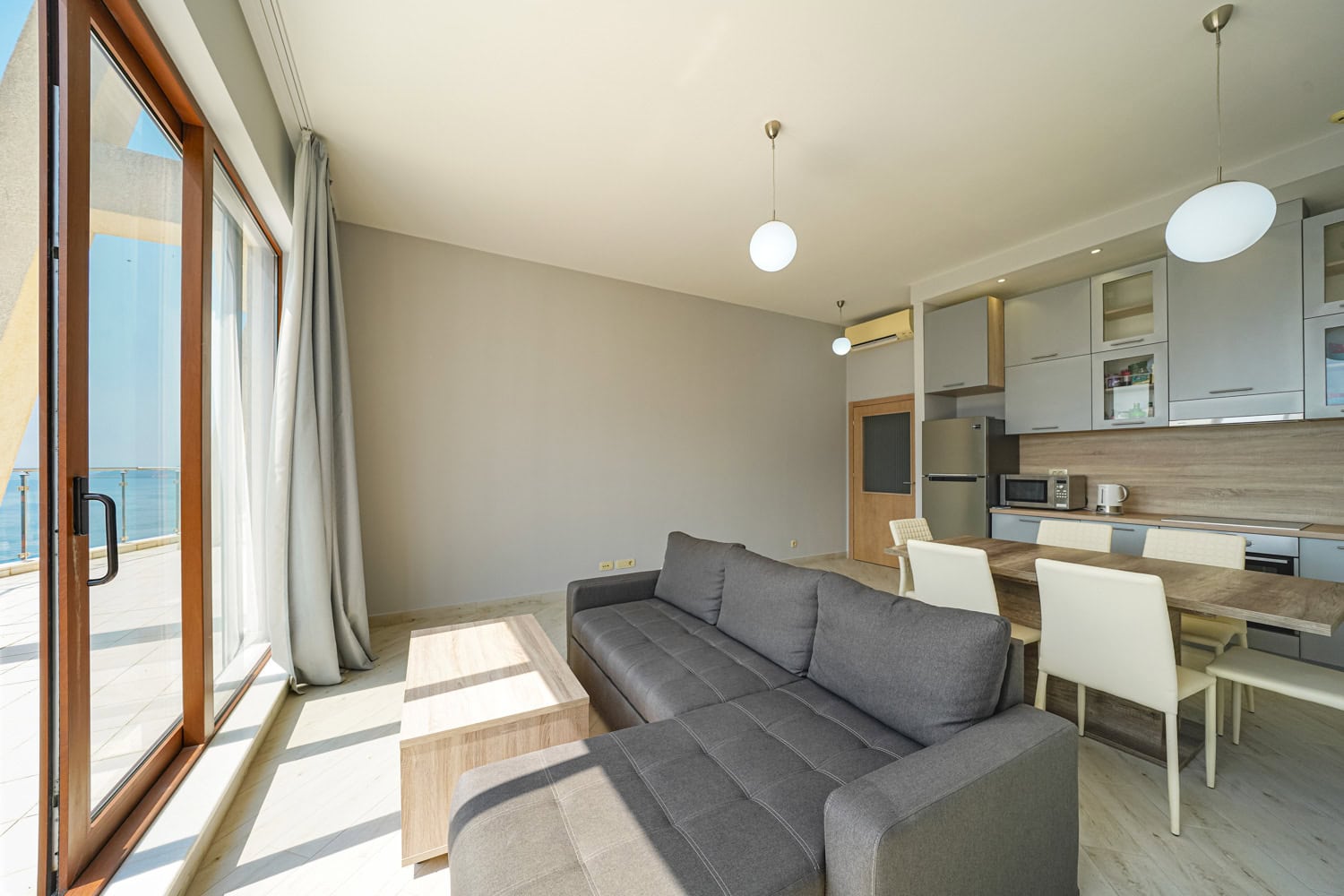 Living room with kitchen in an apartment in Bečići, Montenegro: gray sofa, dining area, and panoramic sea view from the terrace.