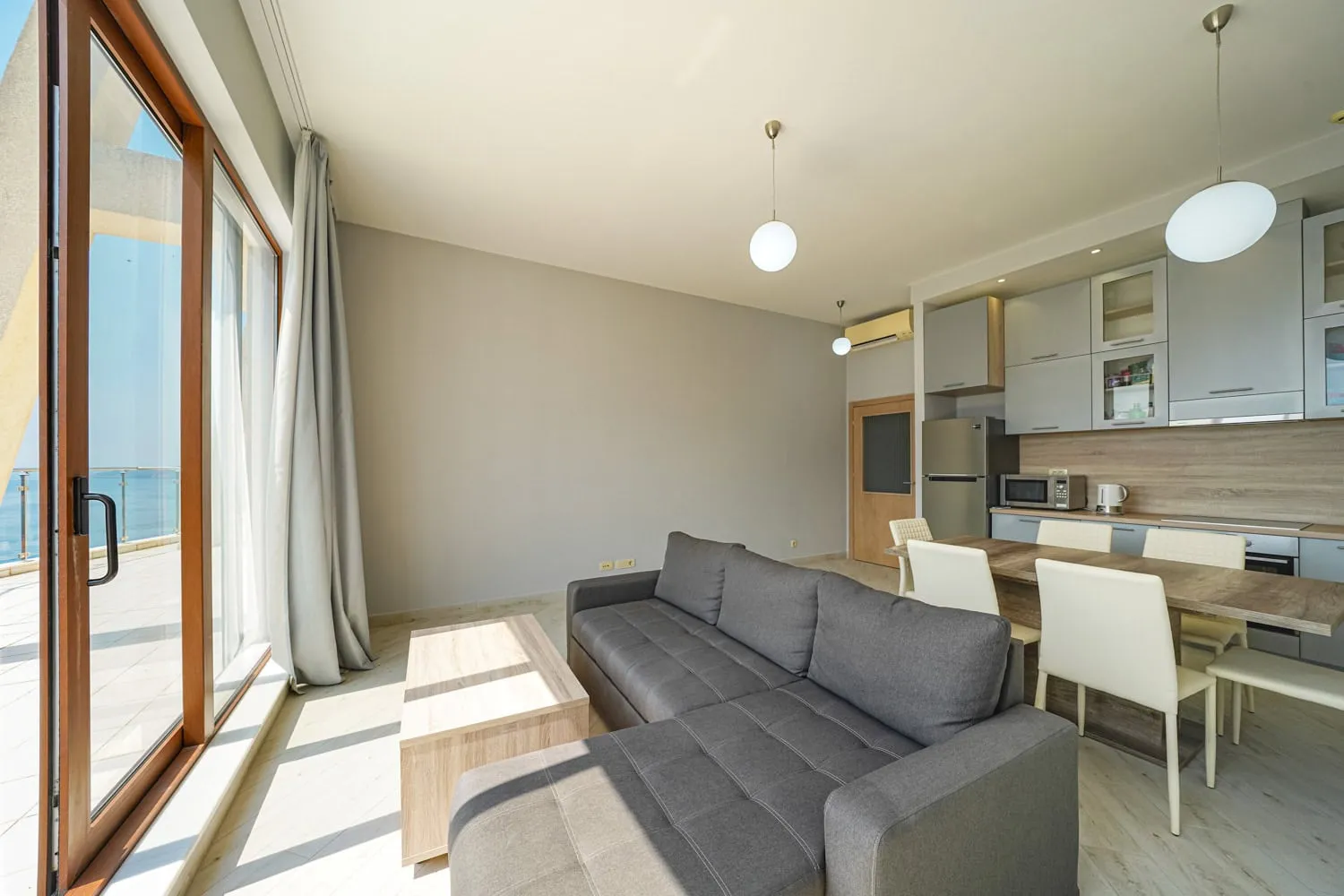 Living room with grey sofa, wooden dining table, and open kitchen area visible near large window.