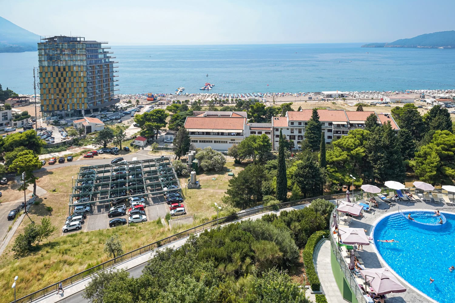 View of the sea, beach, and swimming pool from the apartment terrace in Becici, Montenegro. Panorama of the resort with hotels and parking.