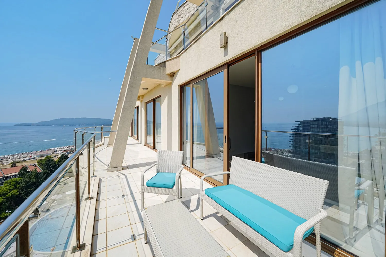 Wide balcony with white wicker furniture and turquoise cushions overlooking the sea and beach.