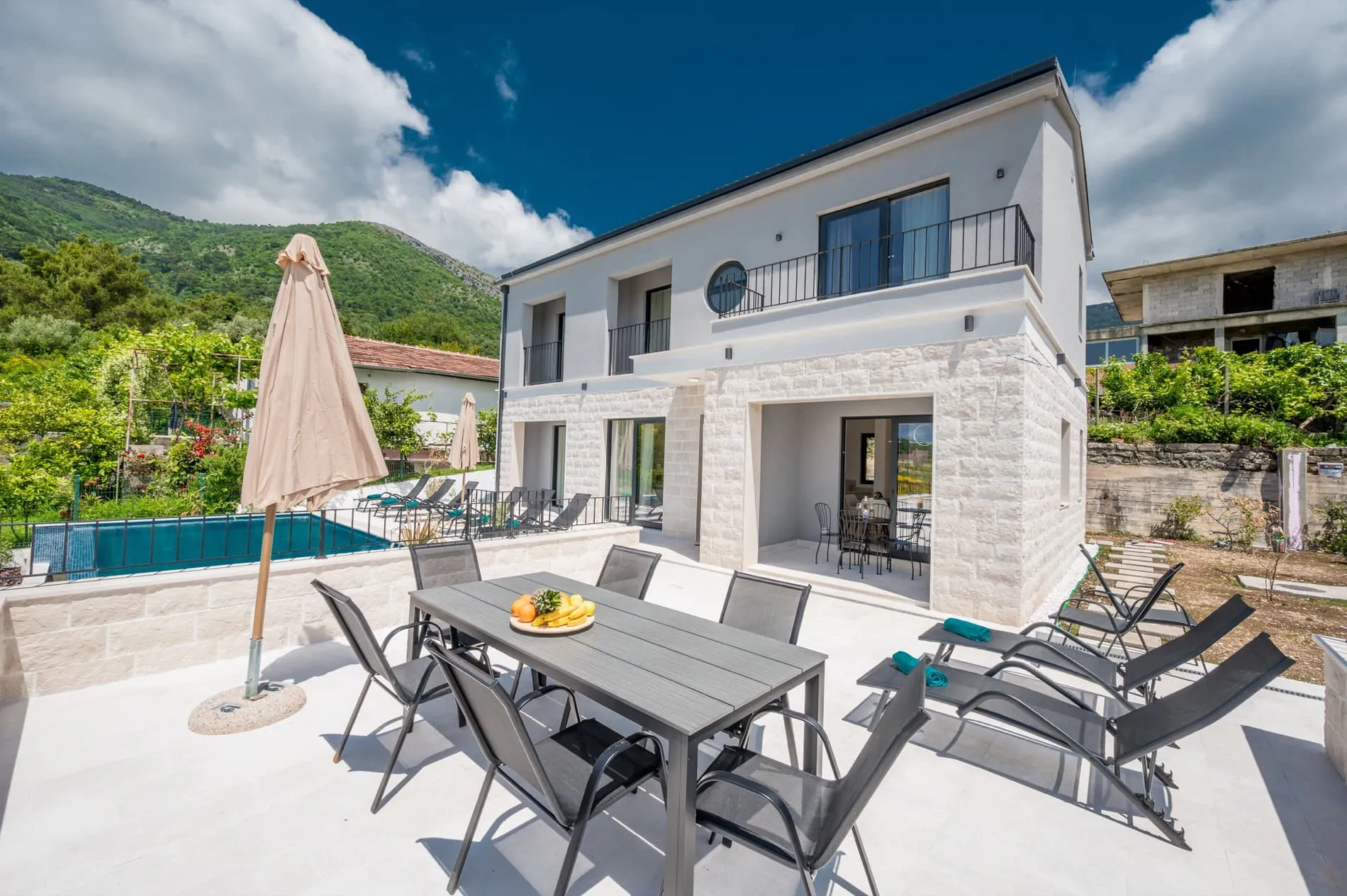 View of a villa facade with a patio, outdoor dining table, sun loungers, and a swimming pool in the background.