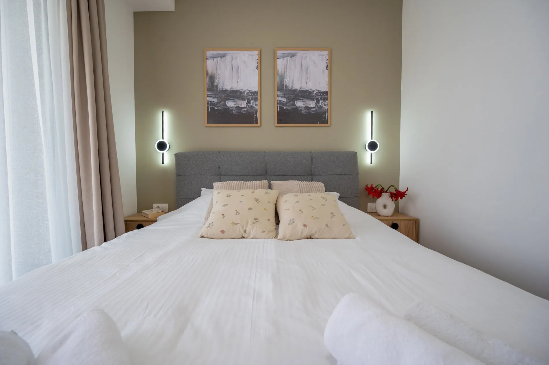 Modern bedroom featuring a large bed with a grey headboard, wall sconces, and abstract artwork.