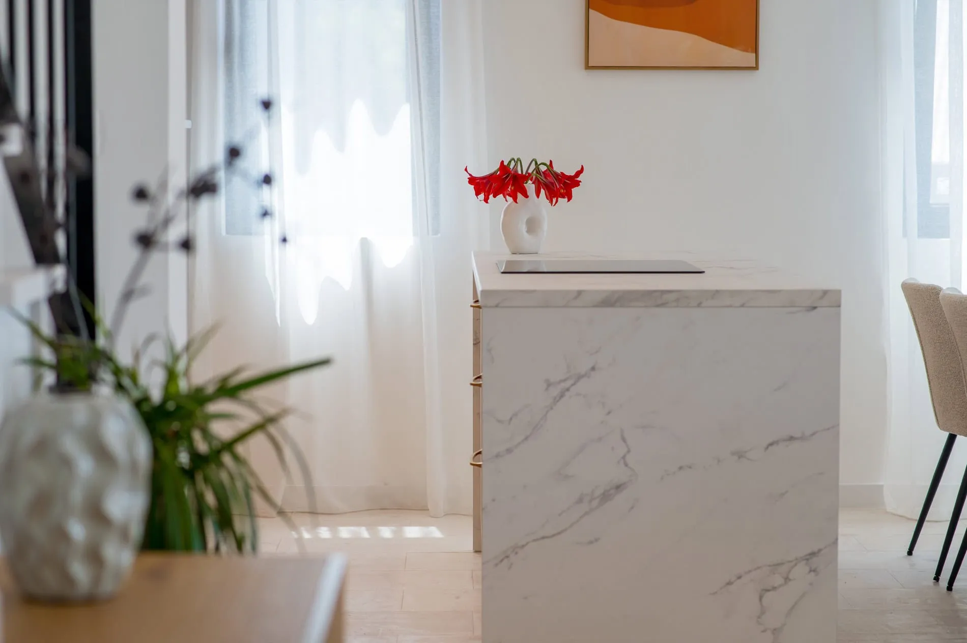 White marble kitchen island with a built-in cooktop and red flowers in a vase against a bright window background.