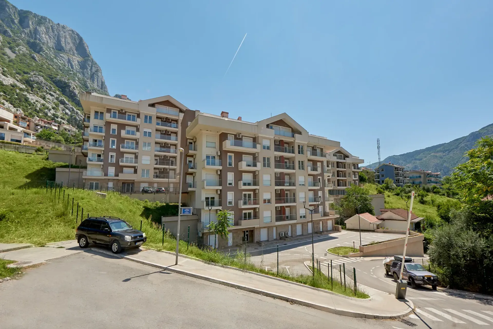 Exterior view of a multi-story apartment complex with balconies and parking against a mountain backdrop.