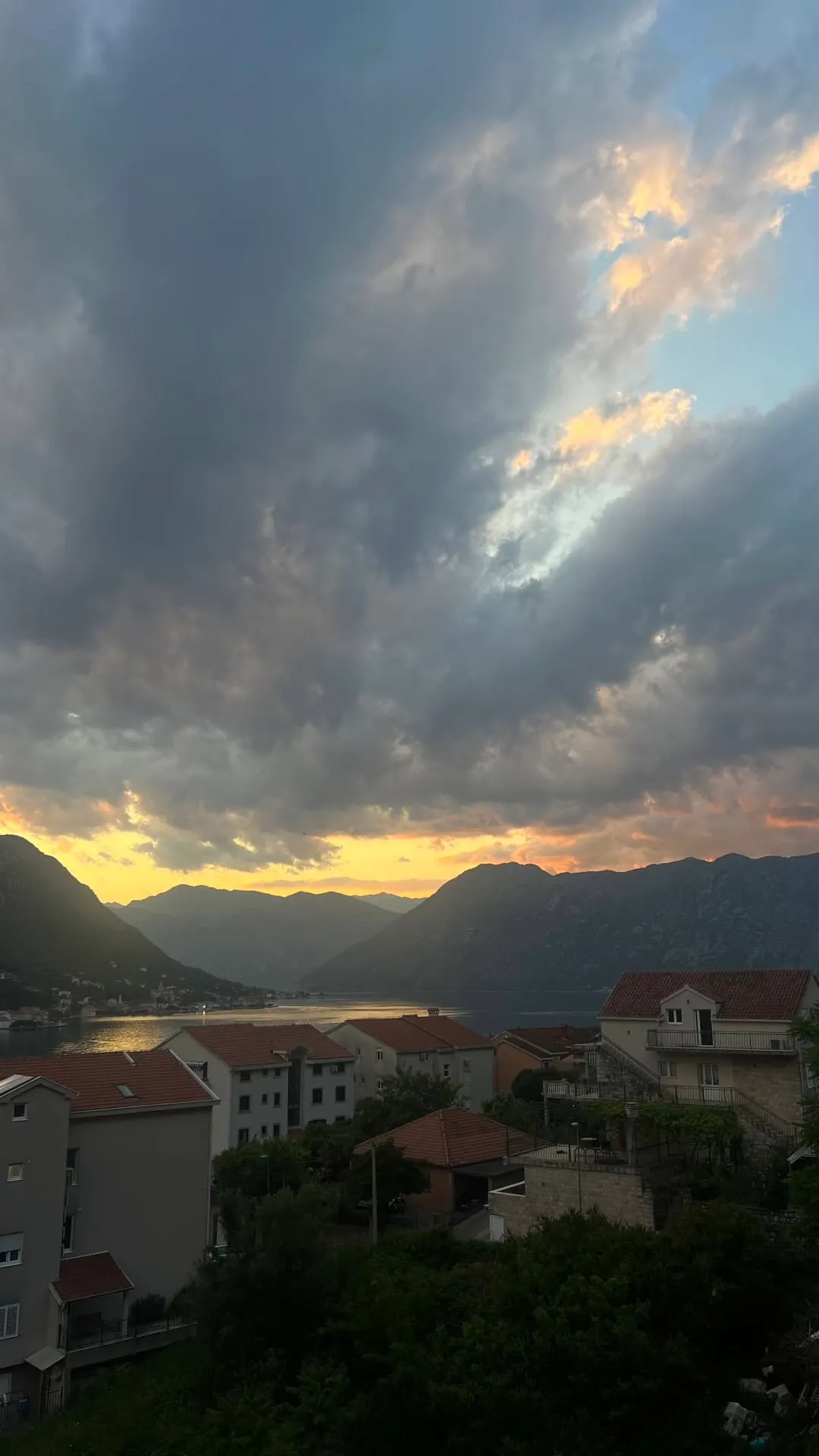 Panoramic view of a bay, mountains, and rooftops under a dramatic cloudy sky at dusk.