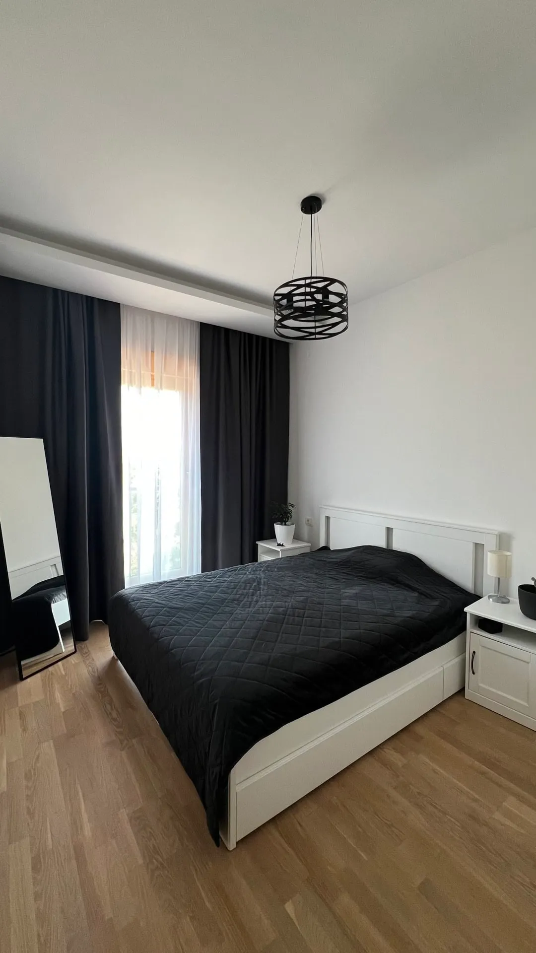 Modern bedroom featuring a white bed frame, black quilted cover, light wood flooring, and a black pendant lamp.