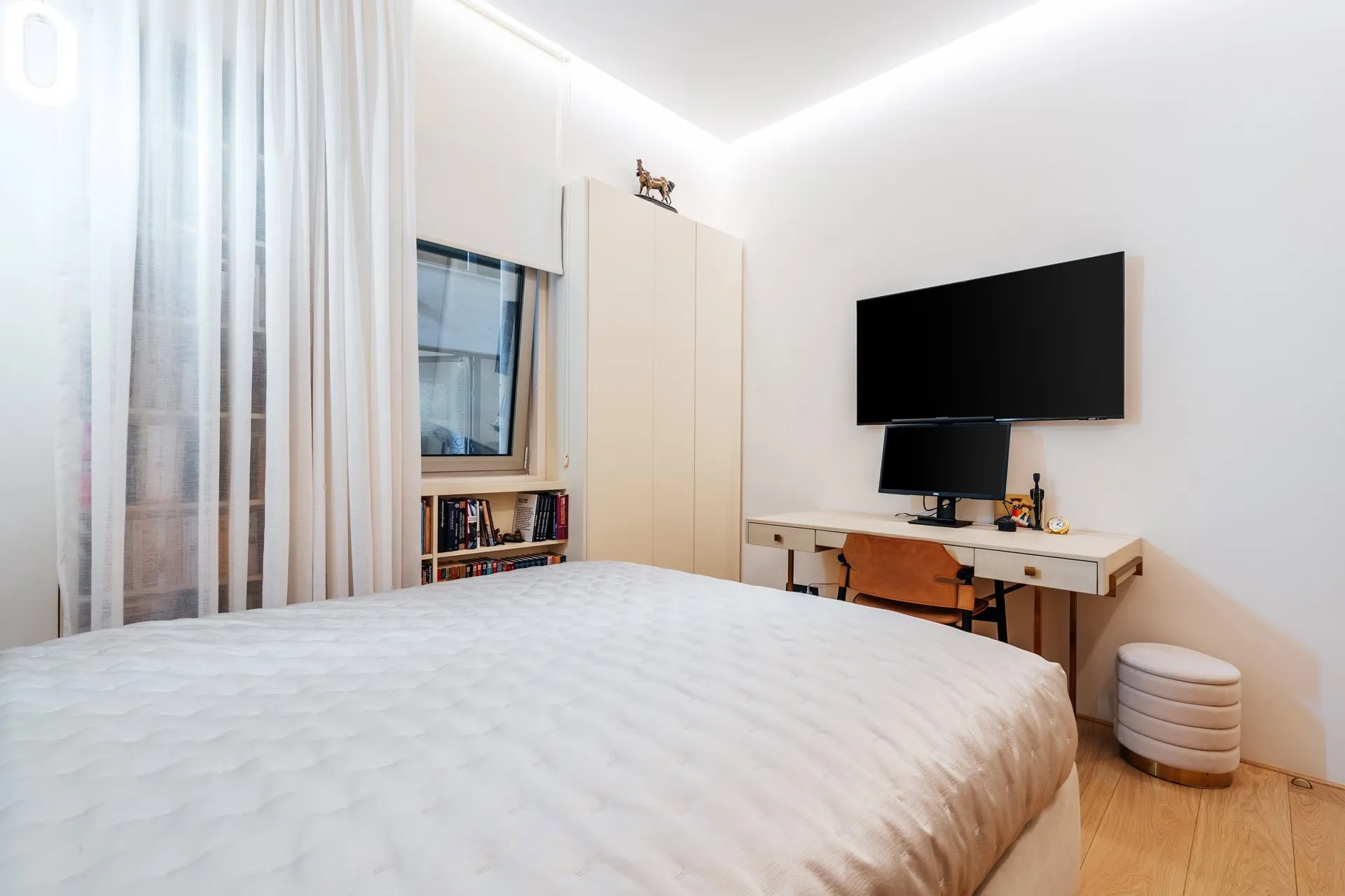Bright bedroom featuring a white bed, desk with TV, wardrobe, and window with sheer curtains.