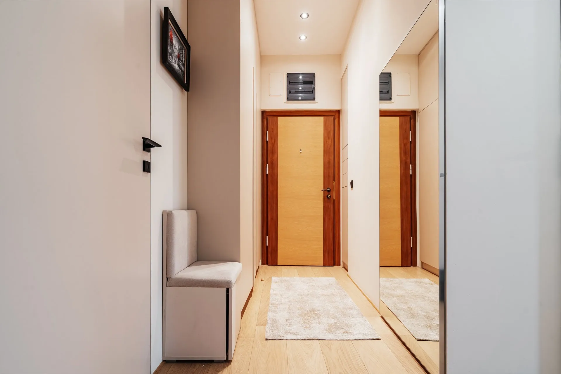 Narrow hallway with a wooden entrance door, upholstered storage bench, light rug, and a large wall mirror reflecting the space.