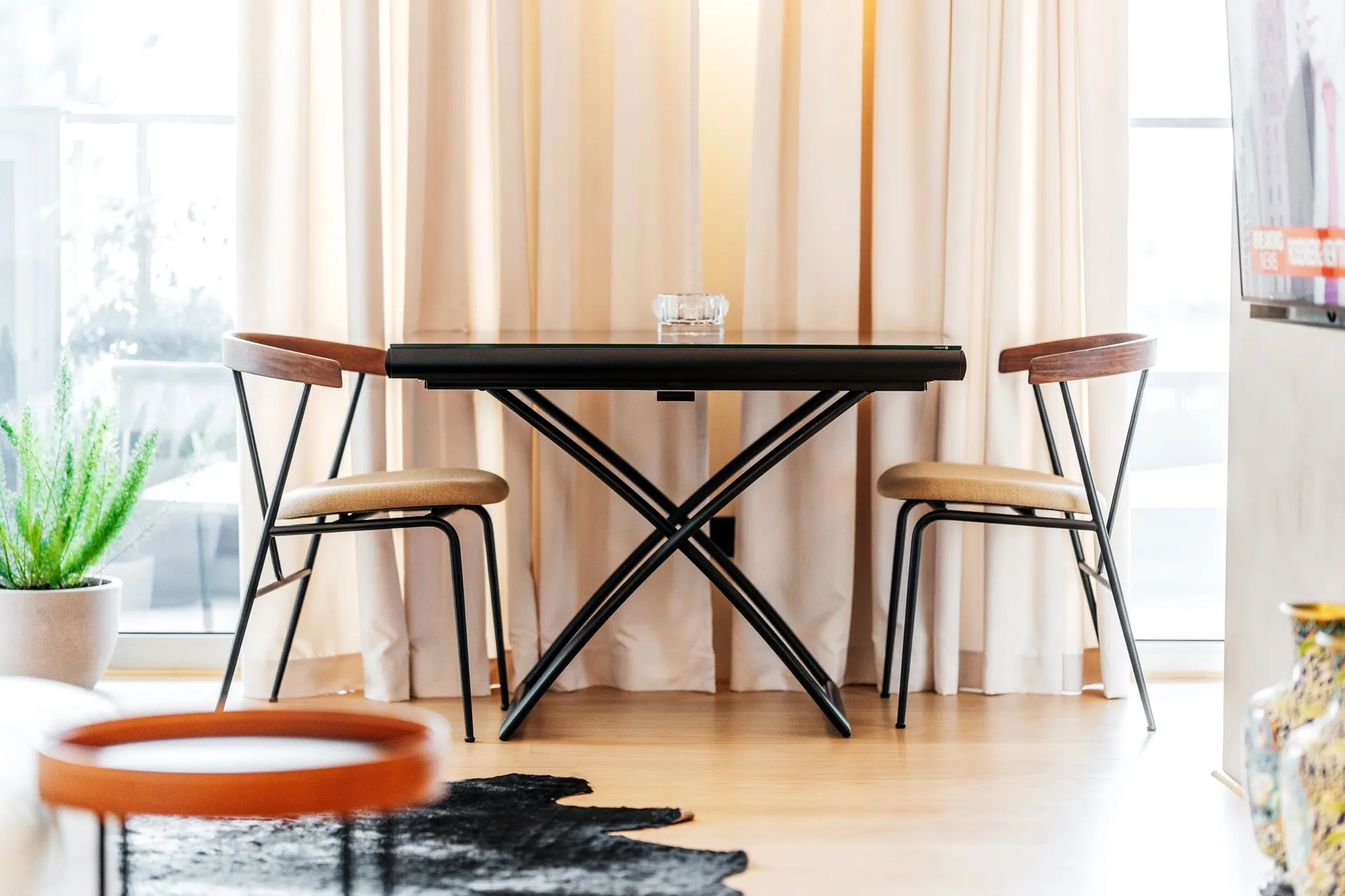 Modern dining area with a black table and two chairs near a window with light curtains and a rug.