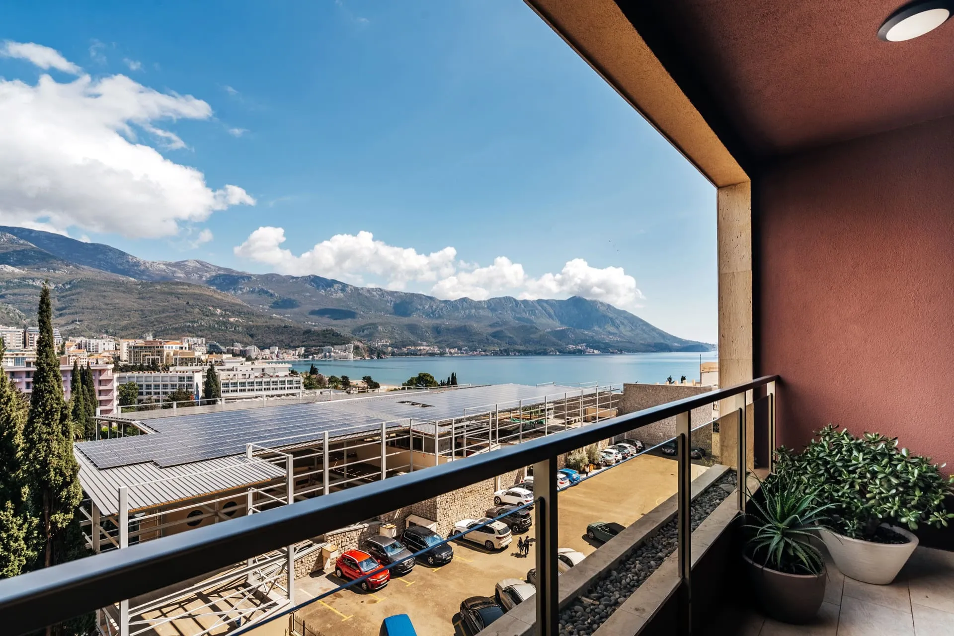 Balcony view overlooking the sea and mountains. Visible metal railing, red wall, and potted plants on tiled floor.