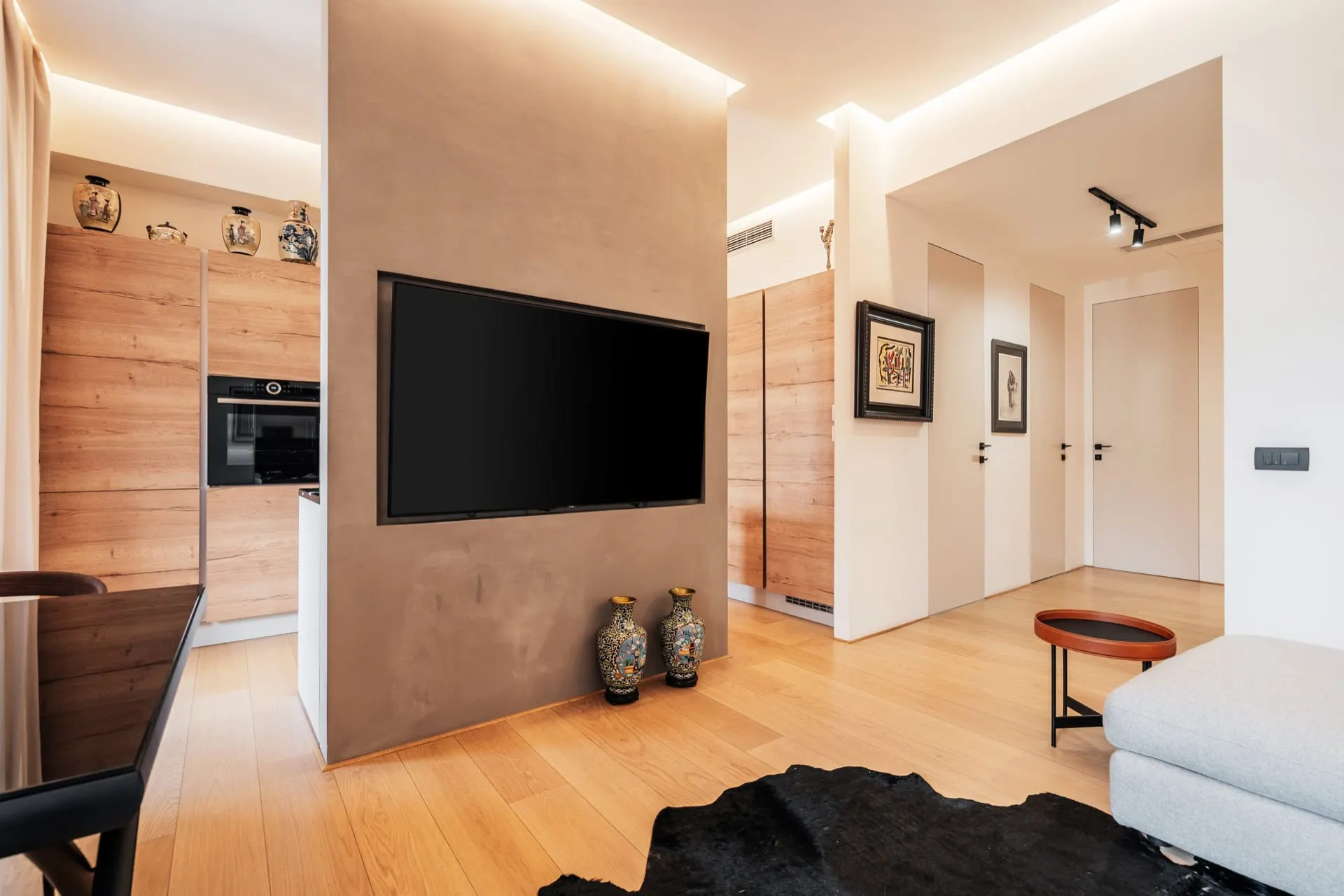 Modern living room with TV on concrete wall, wood floors, and visible kitchen area to the left.