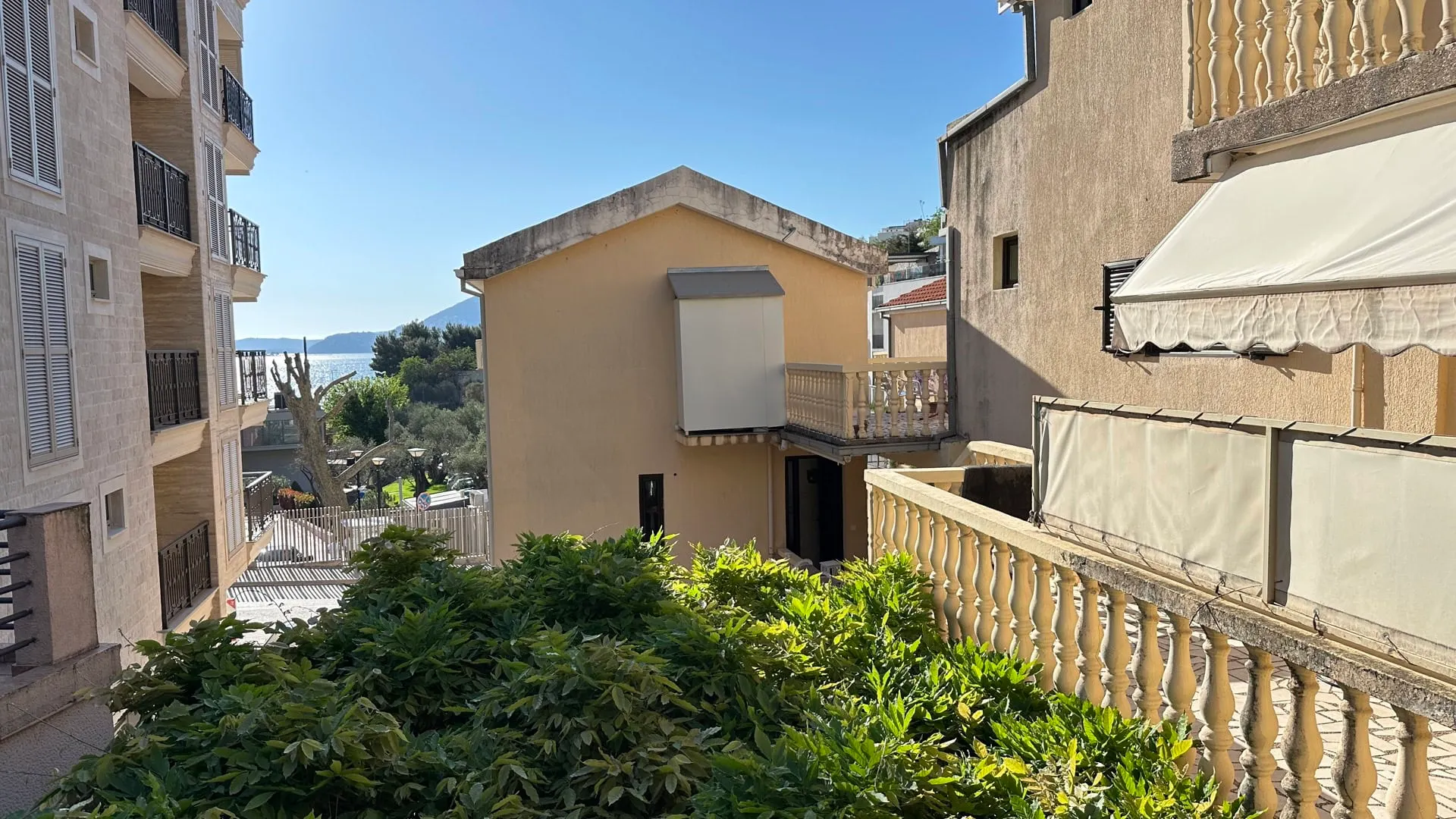 View of buildings with balconies, greenery, and sea in background. Stone railings and awnings.