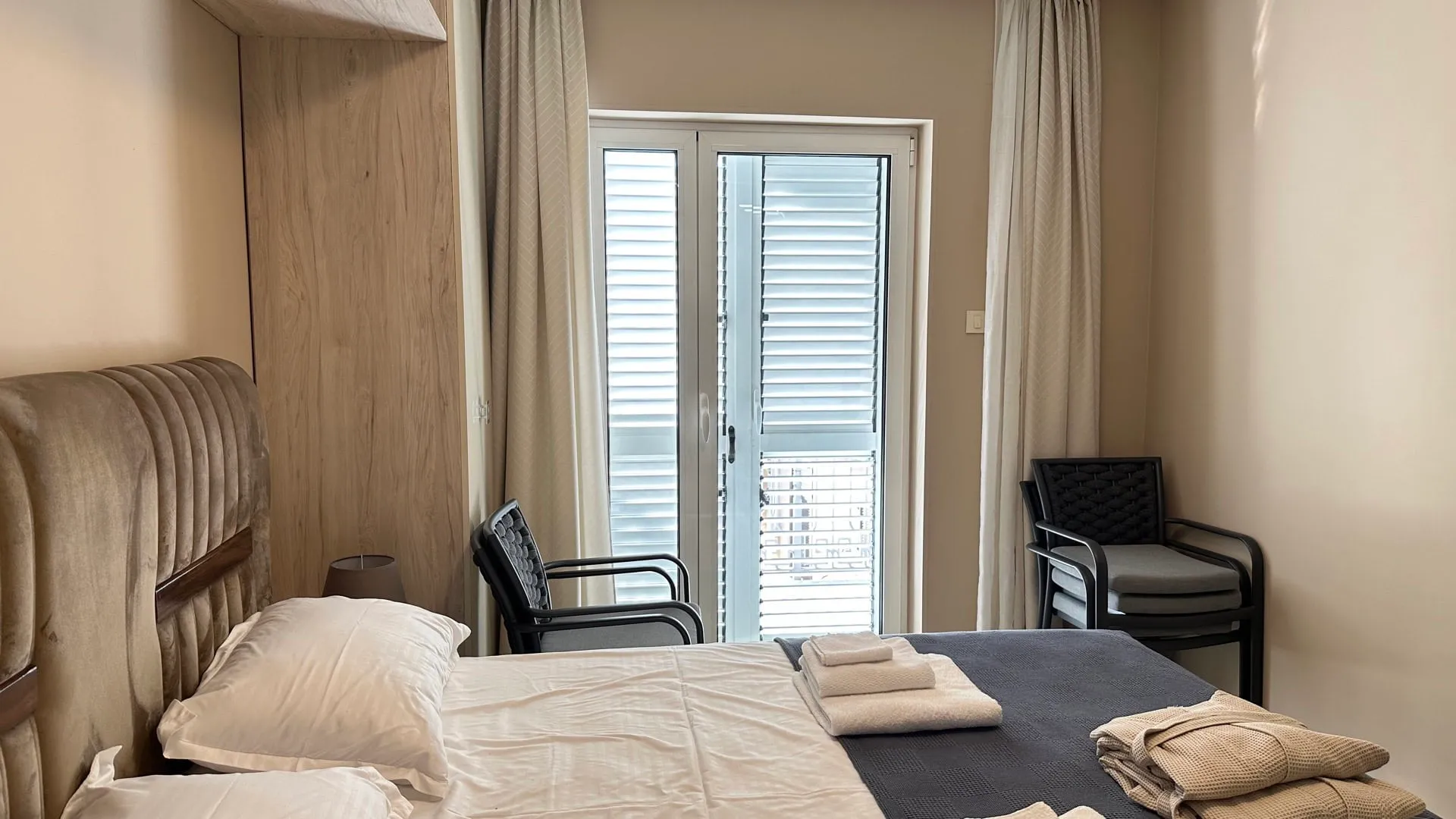 Bedroom featuring a double bed with upholstered headboard, black chairs, and a glass door leading to a balcony.