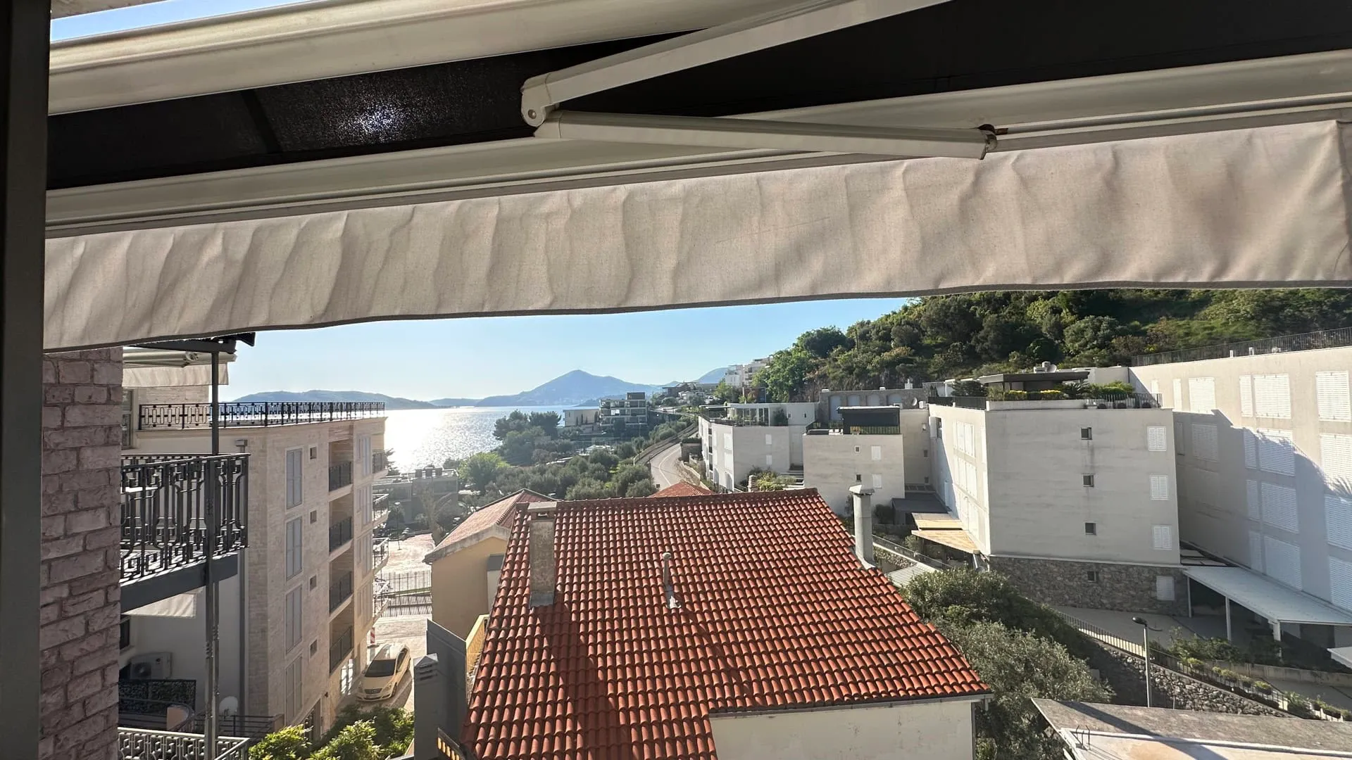 View from a balcony under an awning showing rooftops, sea, and mountains on a sunny day.