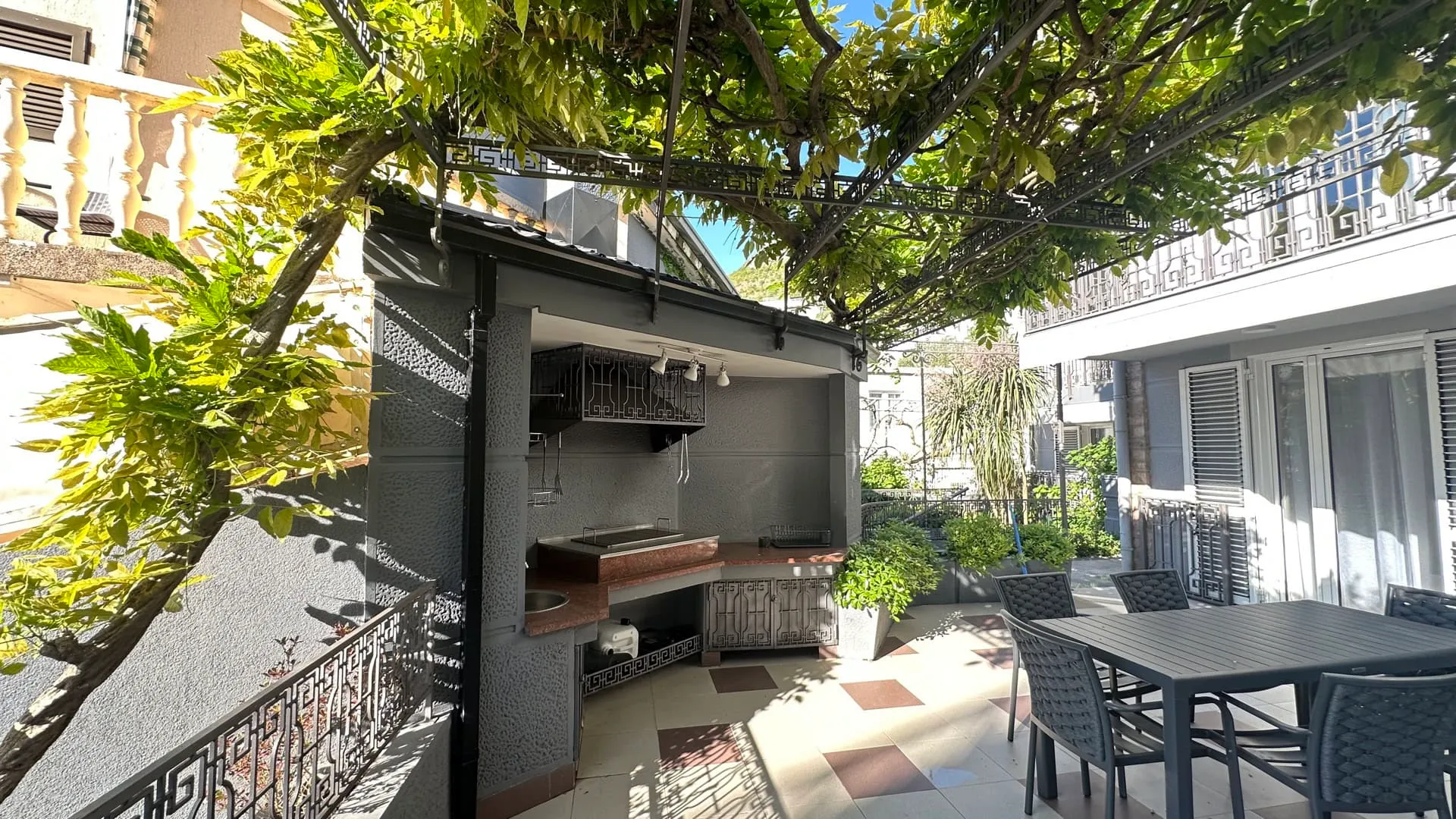 Open terrace featuring an outdoor kitchen, dining set, and a pergola covered with climbing vines.
