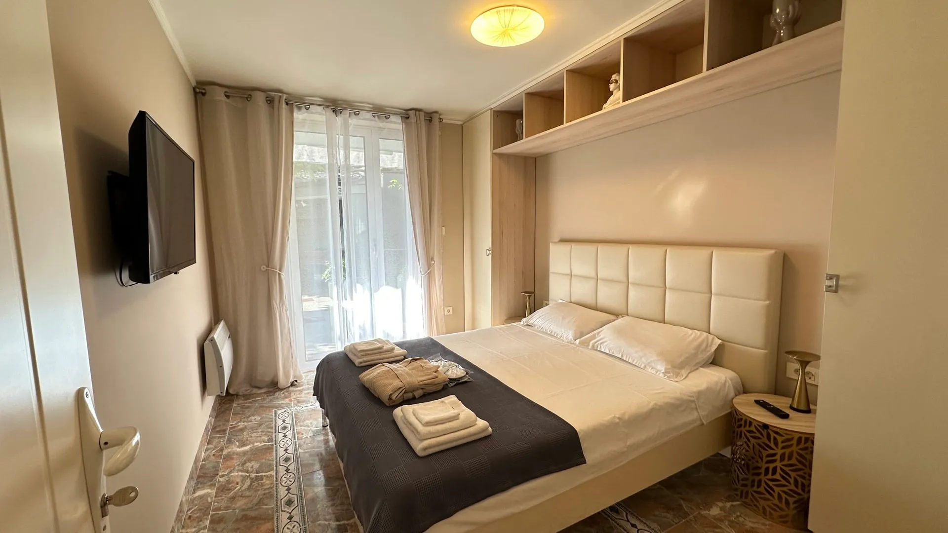 Bedroom featuring a double bed with white linens, wall-mounted TV, built-in wooden shelving, and tiled flooring.