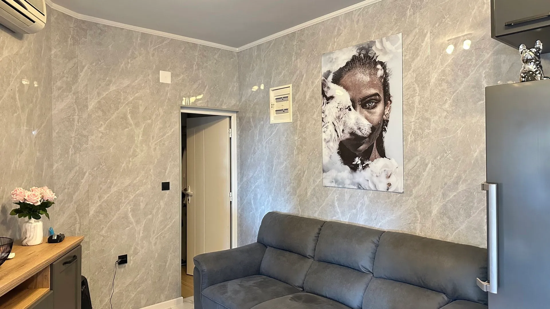Gray sofa, marble walls, large portrait poster, wooden table with roses, and a refrigerator visible on the right.
