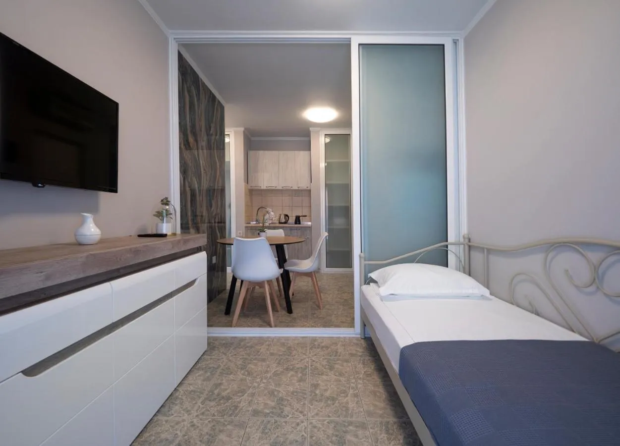 Small bedroom with a single bed, white dresser, and wall-mounted TV; kitchen visible through an open doorway.