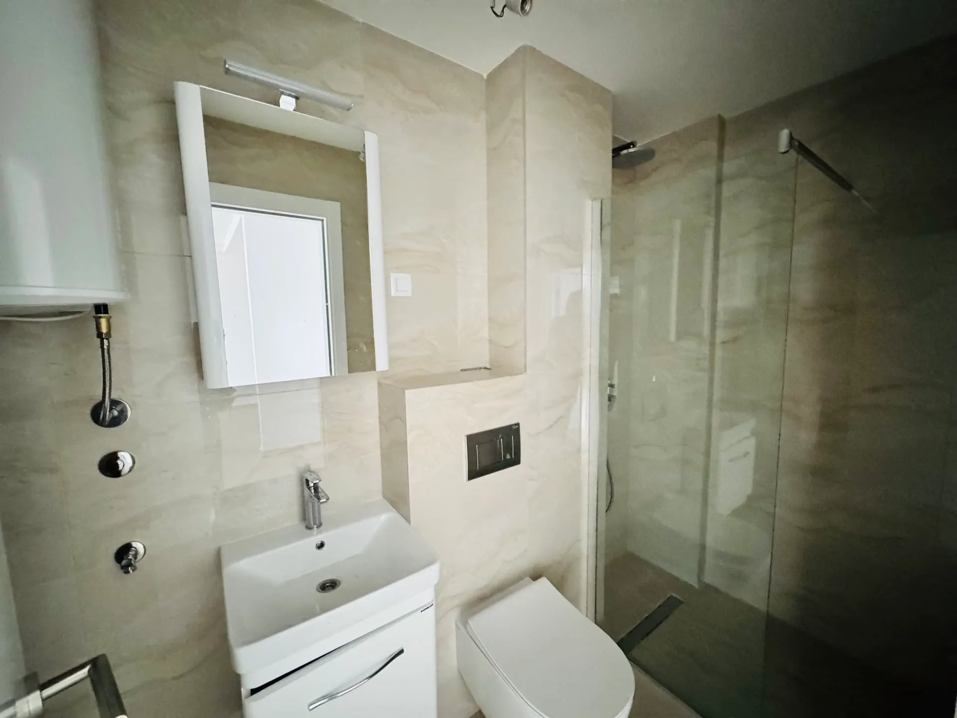 Modern bathroom featuring a white vanity sink, wall-mounted toilet, glass shower enclosure, and beige marble-style tiles.
