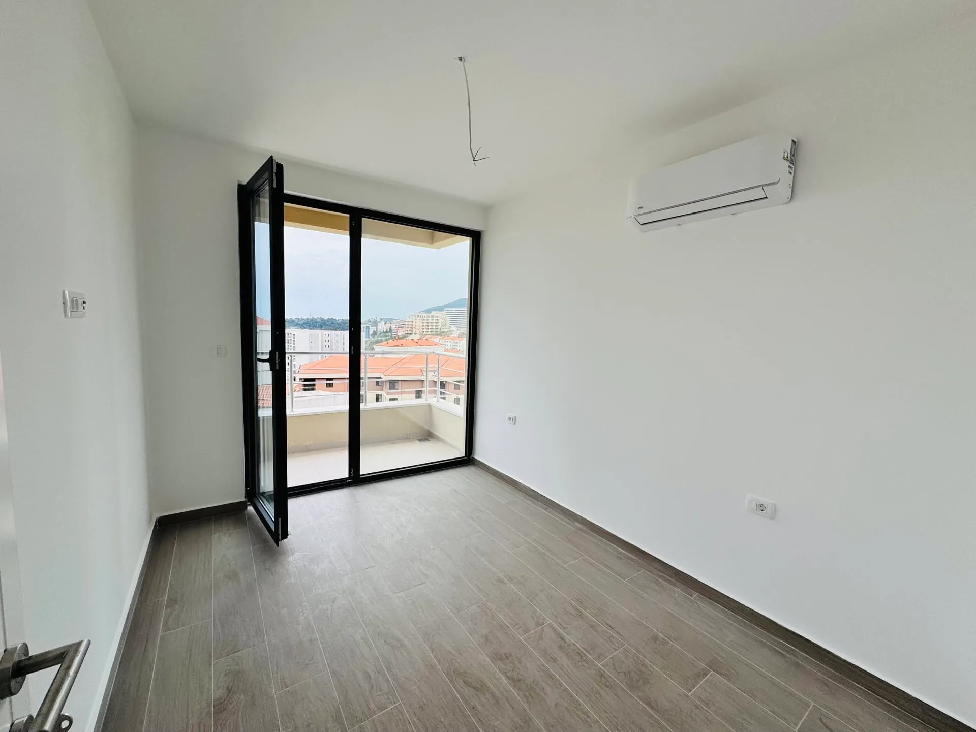 Empty room with grey wood-look tiles, white walls, wall-mounted AC unit, and balcony access.