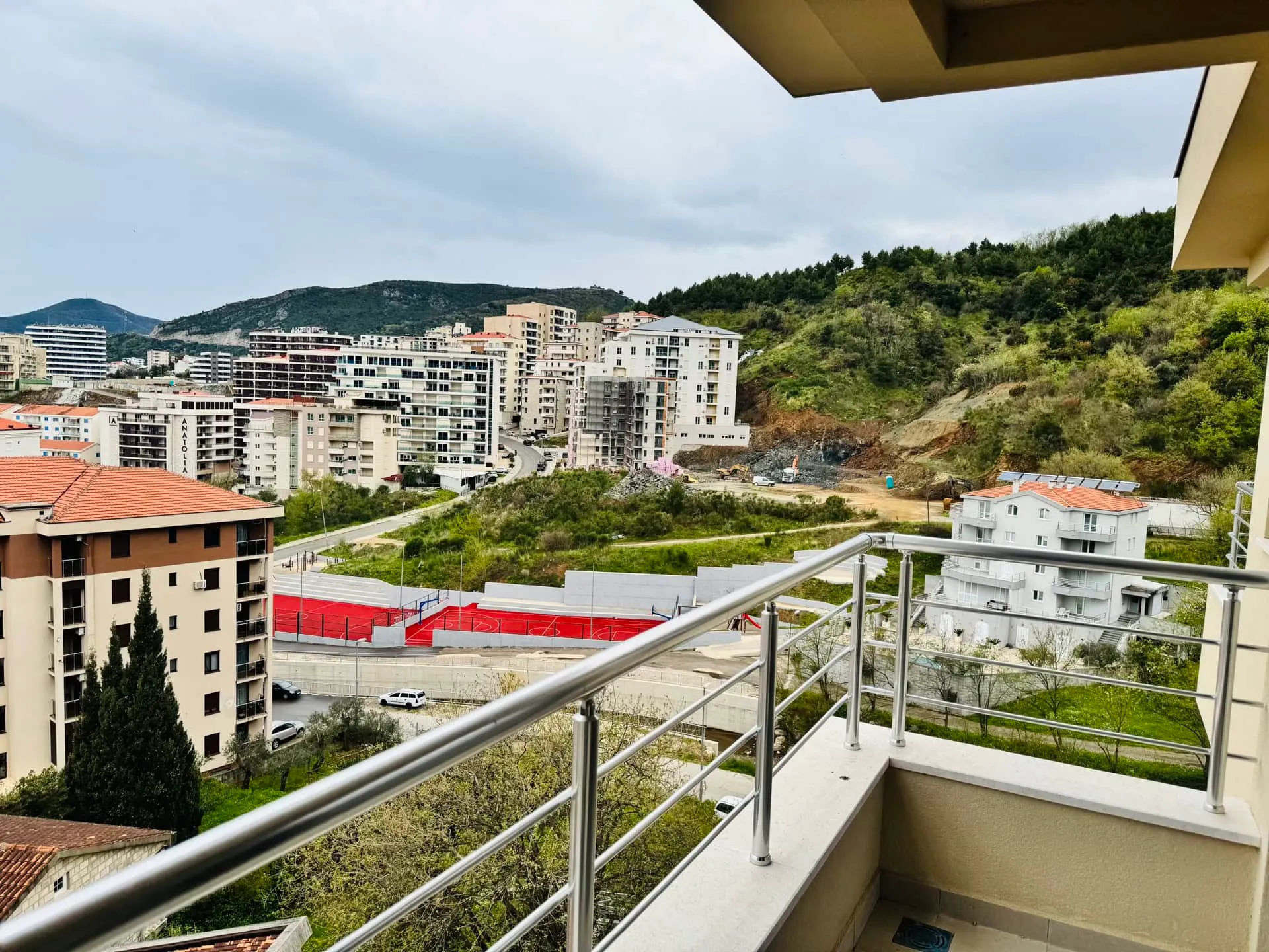 View from a balcony with metal railings overlooking a city, red sports court, and green hills.
