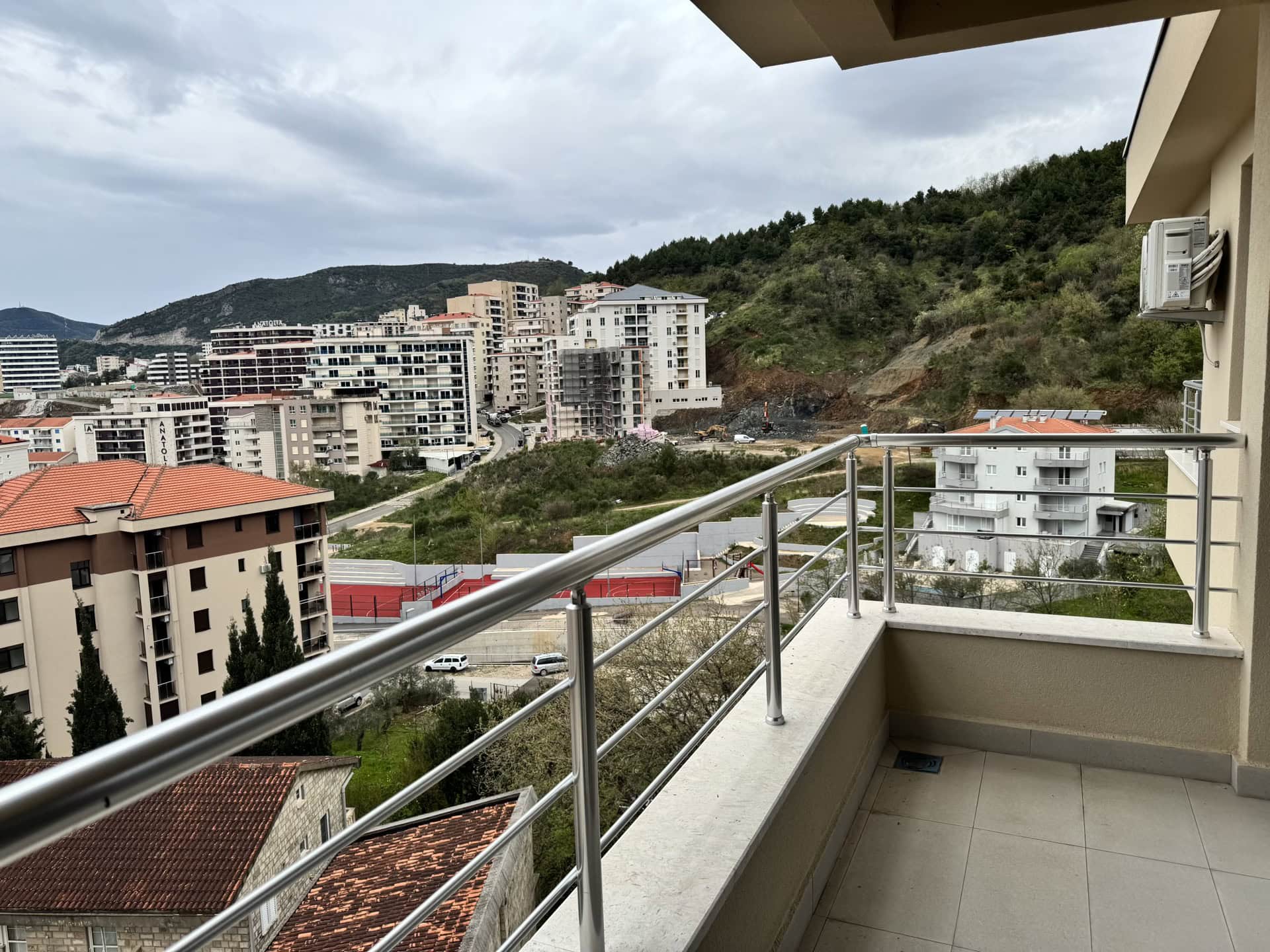View from the apartment balcony in Bečići, Montenegro: a city panorama, modern residential complexes, and mountains in the background.