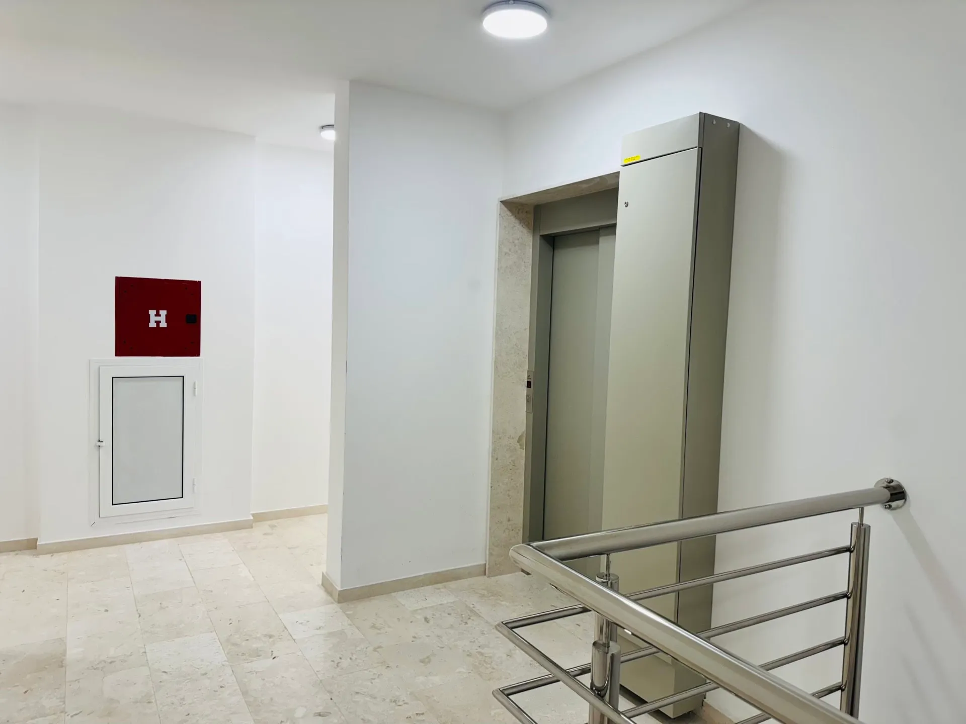 Elevator lobby with metal railing, white walls, tiled floor, and a red fire safety panel on the wall.