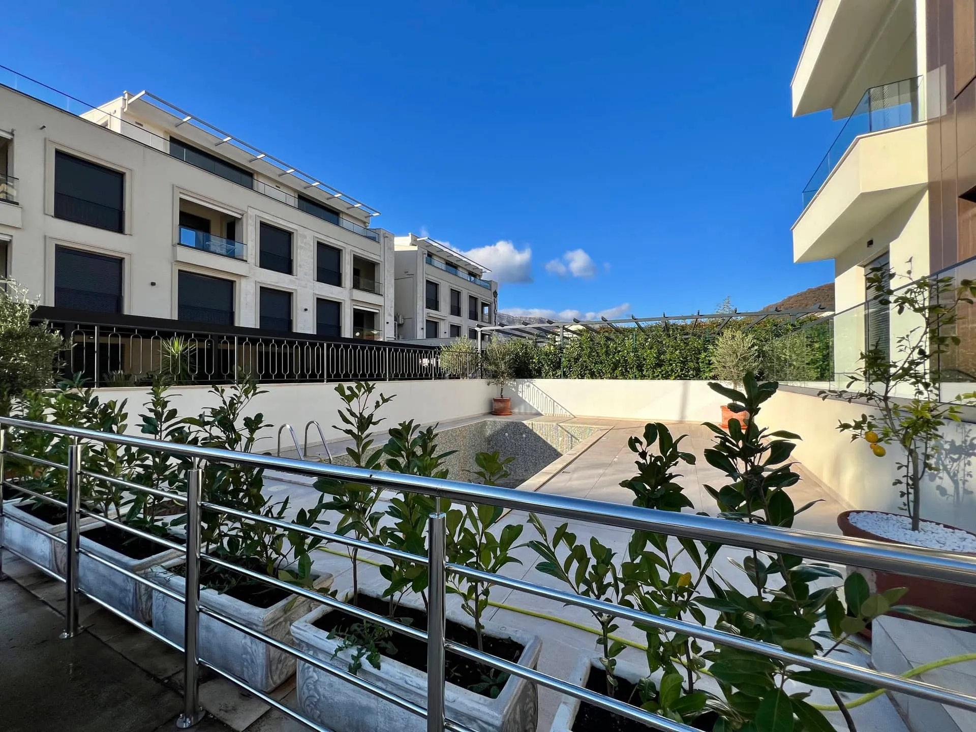 Open pool with metal ladder surrounded by potted plants in stone beds and modern residential buildings under blue sky.